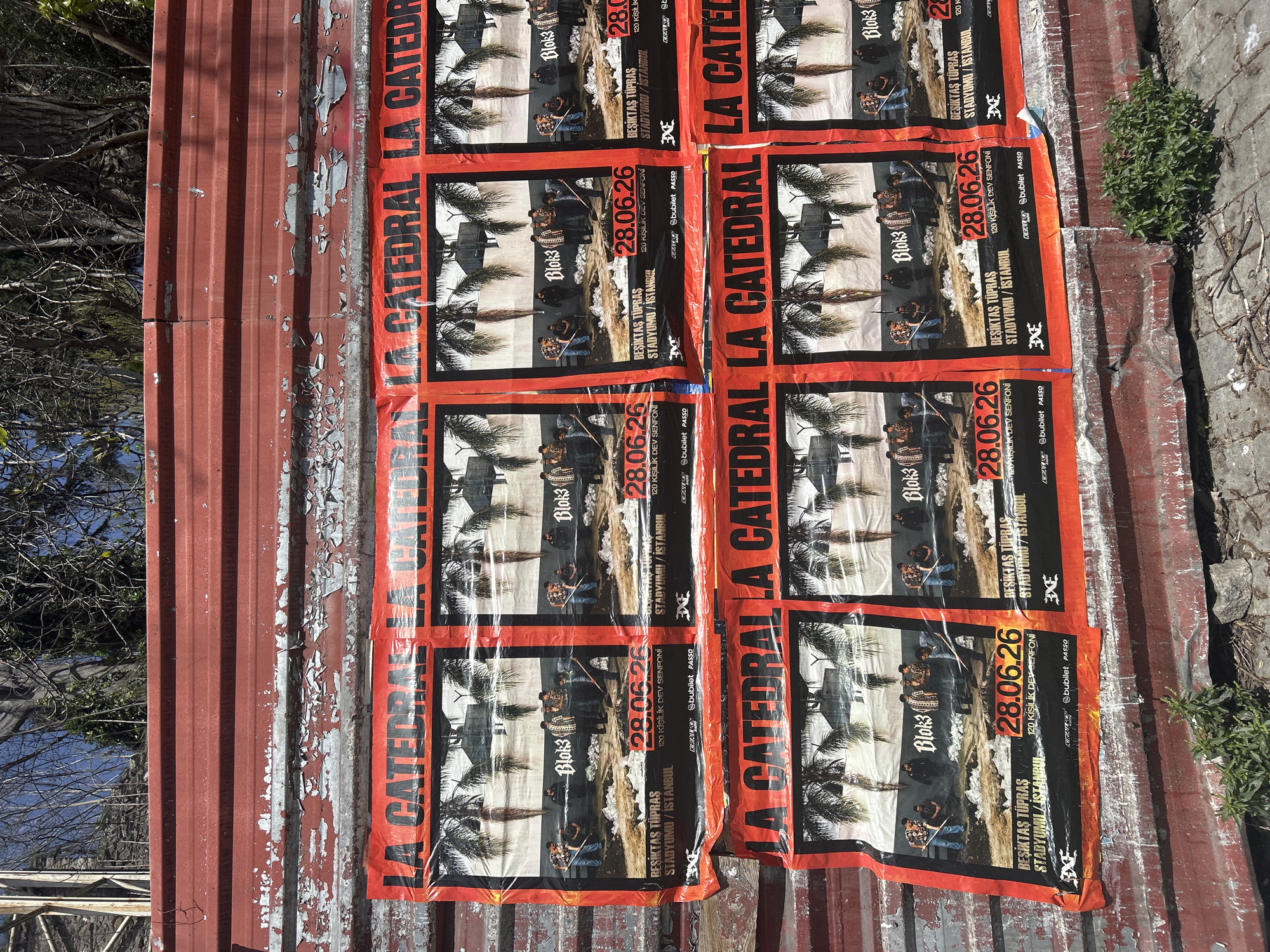 The image shows a series of posters adhered to a corrugated metal surface, likely a wall or roof. The posters are printed with an advertisement for an event called "LA CATEDRAL" featuring "Blok3" on June 28, 2026 at "BEŞİKTAŞ TÜPRAS STADYUMU/İSTANBUL". The posters are orange, black, and white, and the text includes phrases in Turkish. The corrugated metal surface shows signs of aging with flaking paint. The photo is taken during daylight, as evidenced by the shadows and lighting. Vegetation is visible on the right side of the image, and some bare branches of trees appear in the background along with a glimpse of the sky. The location is presumably in Istanbul, due to Turkish text used in the concert announcement. It is difficult to ascertain with certainty if this is Beyoğlu, but based on the subject matter, it is possible.
