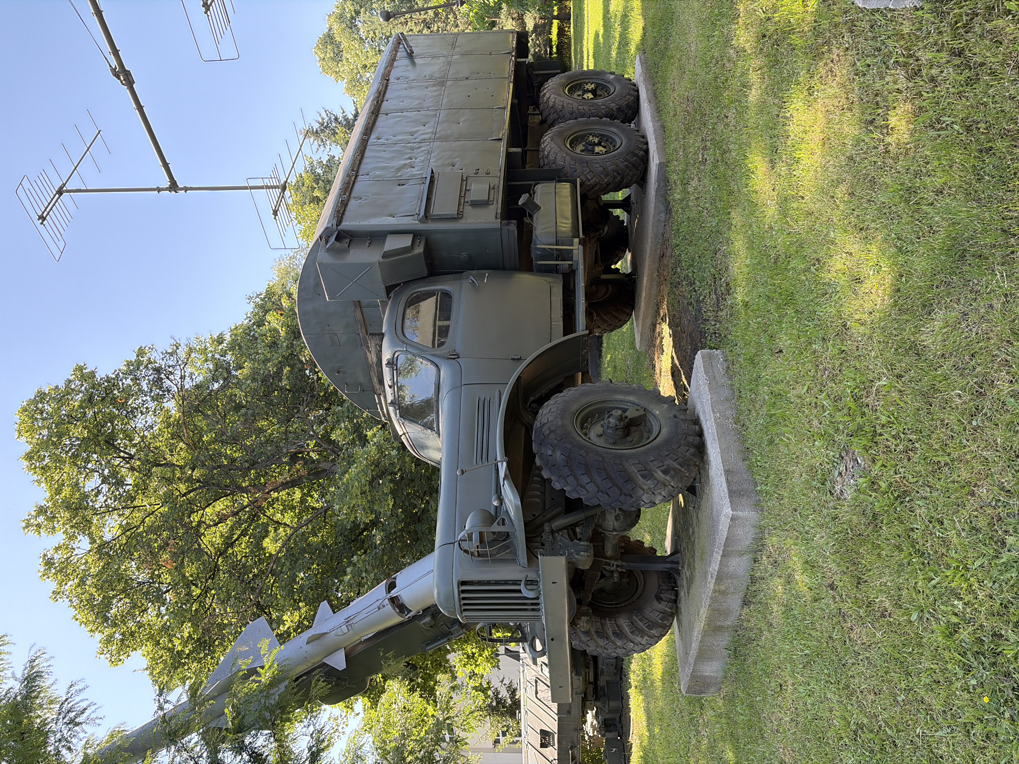 The image displays a vintage military truck, likely a ZIL-131, with a missile launcher mounted on its rear. The truck is painted in a drab green color and features large, rugged tires. It is positioned on a concrete pad in a grassy outdoor area. Behind the truck, a tall antenna array is visible. The scene appears to be set in a park or an open-air museum.
This specific military hardware is on display at the National Museum of Military History in Sofia, Bulgaria. The museum grounds are known for showcasing various military vehicles and equipment, often situated in a park-like setting with trees and open lawns, consistent with the environment depicted.