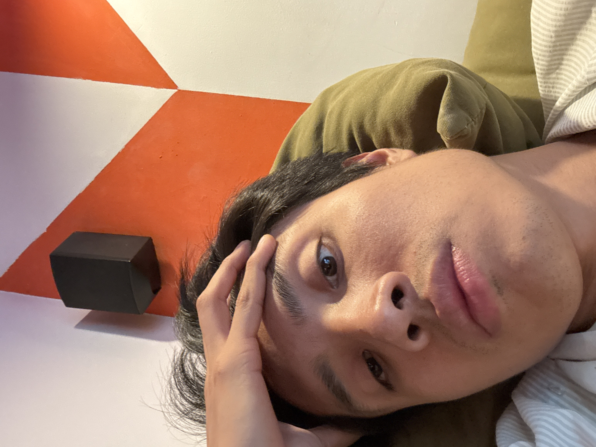 A close-up, rotated portrait captures a person, likely male, lying down indoors. The individual has dark hair and brown eyes, looking directly towards the viewer with a neutral to slightly pensive expression. One hand rests gently on their forehead and hairline.
The person is supported by what appears to be a khaki or olive-green fabric, possibly a pillow or blanket, which is visible behind their head and neck. On the right side of the frame, a striped fabric in light gray and white is visible, likely part of their clothing or another textile.
To the left of the person's head, a small, black rectangular object, possibly a speaker or a decorative cube, is positioned against a wall. The background features a striking geometric pattern of large triangles in contrasting bright orange and white, indicating a modern and visually interesting indoor setting. The photograph is taken in São Paulo, Brazil. No text is visible in the image.