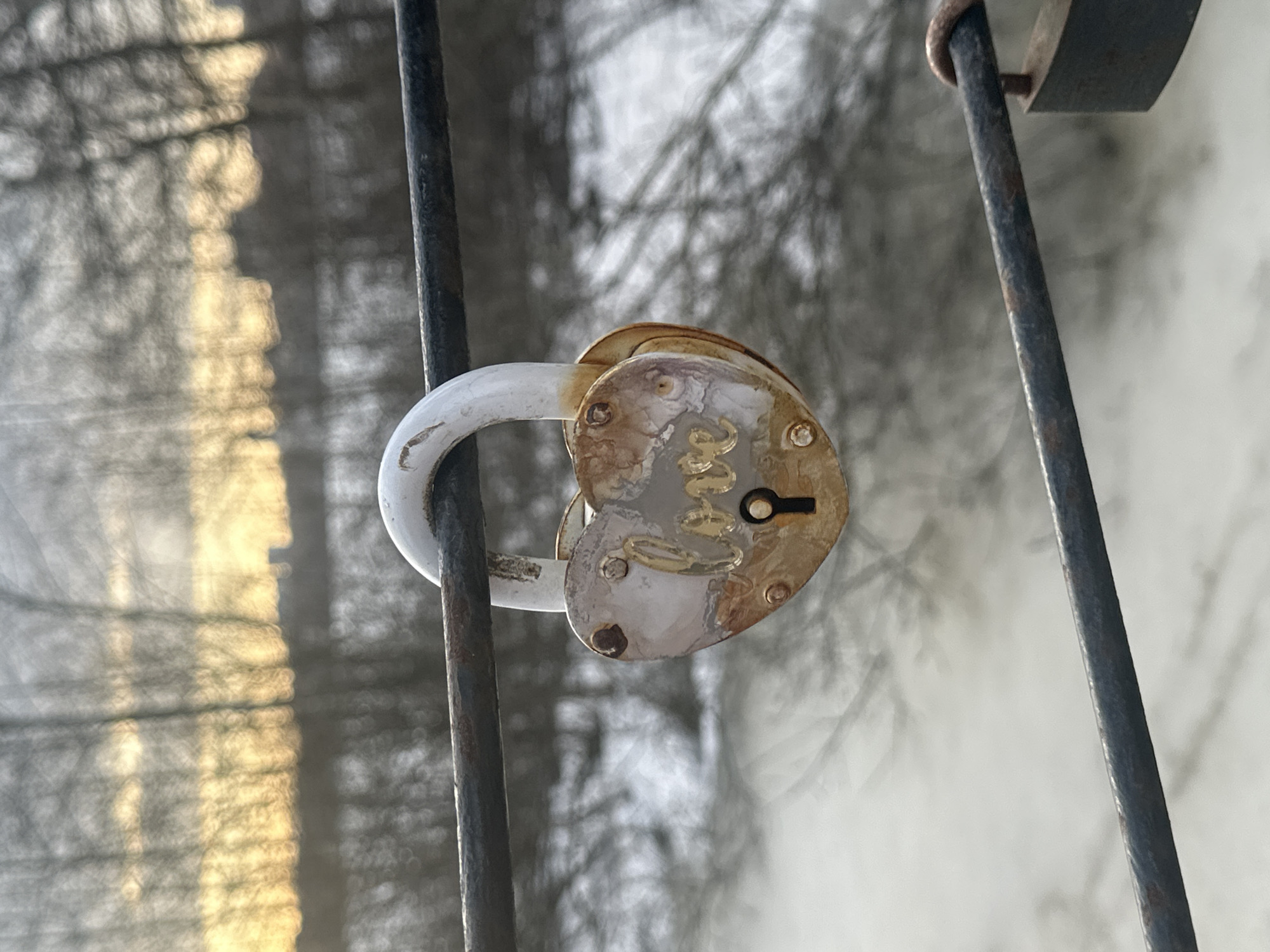 A close-up shot shows a heart-shaped padlock hanging from a dark metal grate. The padlock is painted white, but the paint is peeling and there is visible rust. The lock mechanism is visible, including a keyhole. The background is blurred, but appears to be a wintery scene with bare trees. The setting is the Tretyakov Bridge in Moscow, Russia, known for love locks attached to its railings.