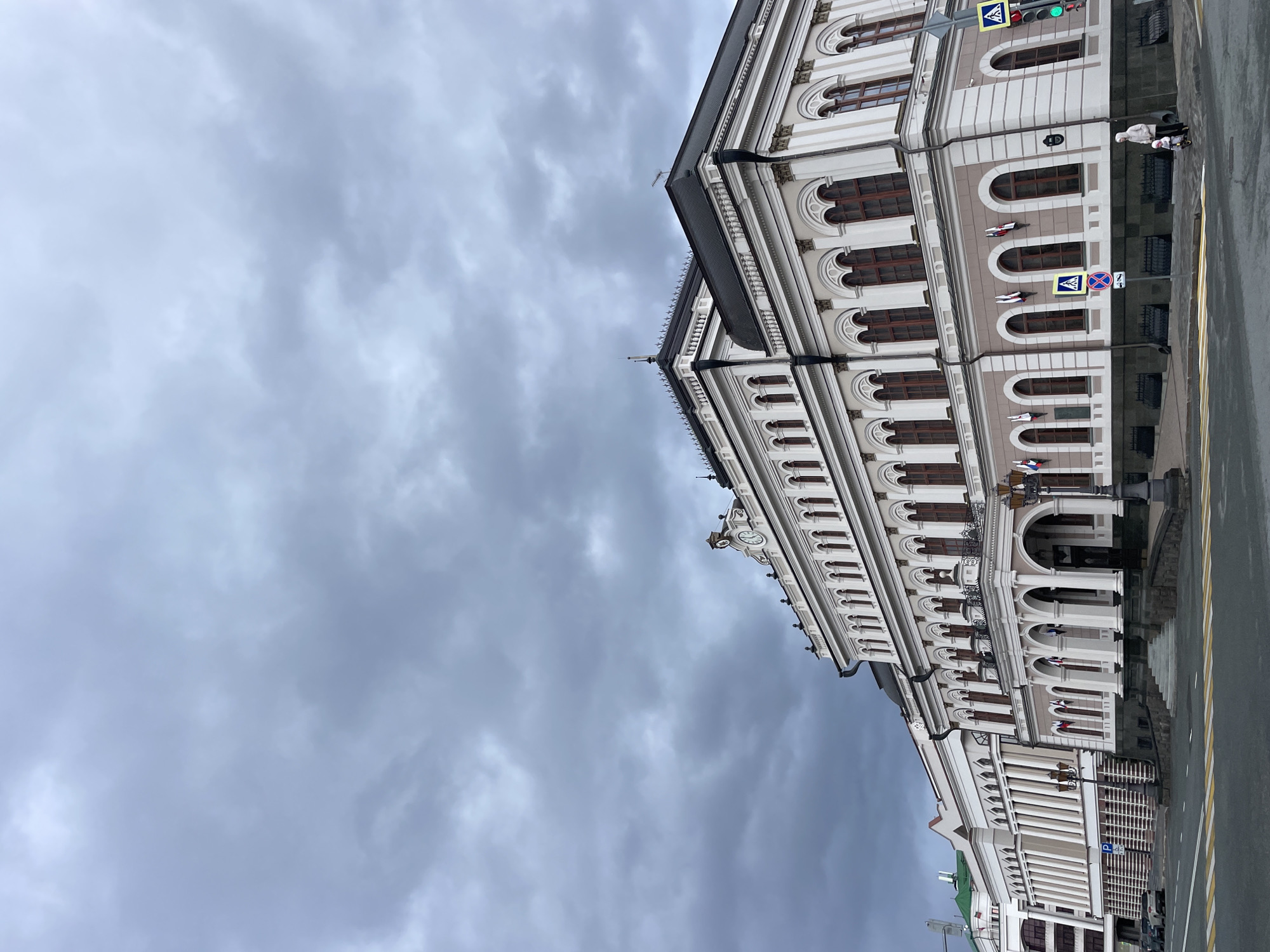 The image displays a grand, multi-story building with elaborate classical architecture under an overcast sky. The building's facade is light cream or beige, featuring numerous arched windows with dark frames and decorative elements like columns, cornices, and intricate moldings. Small banners or flags in red, white, and blue are visible hanging from some windows, and a clock face is set into an upper section of the building.
A wide, grey, cloudy sky dominates a large portion of the frame, indicating an overcast day. At the street level, a wet paved road with yellow markings is visible, alongside a sidewalk that reflects the building and sky. Two individuals, one pushing a stroller, are seen walking on the sidewalk, suggesting a typical urban scene.
Street furniture includes a traffic light displaying a green signal, and several road signs: a blue pedestrian crossing sign, a "No Stopping" sign (red circle with a blue background and red diagonal line), and a blue parking sign with a white 'P'. The overall scene depicts a bustling street in Kazan, Russia, during what appears to be a damp, grey day.