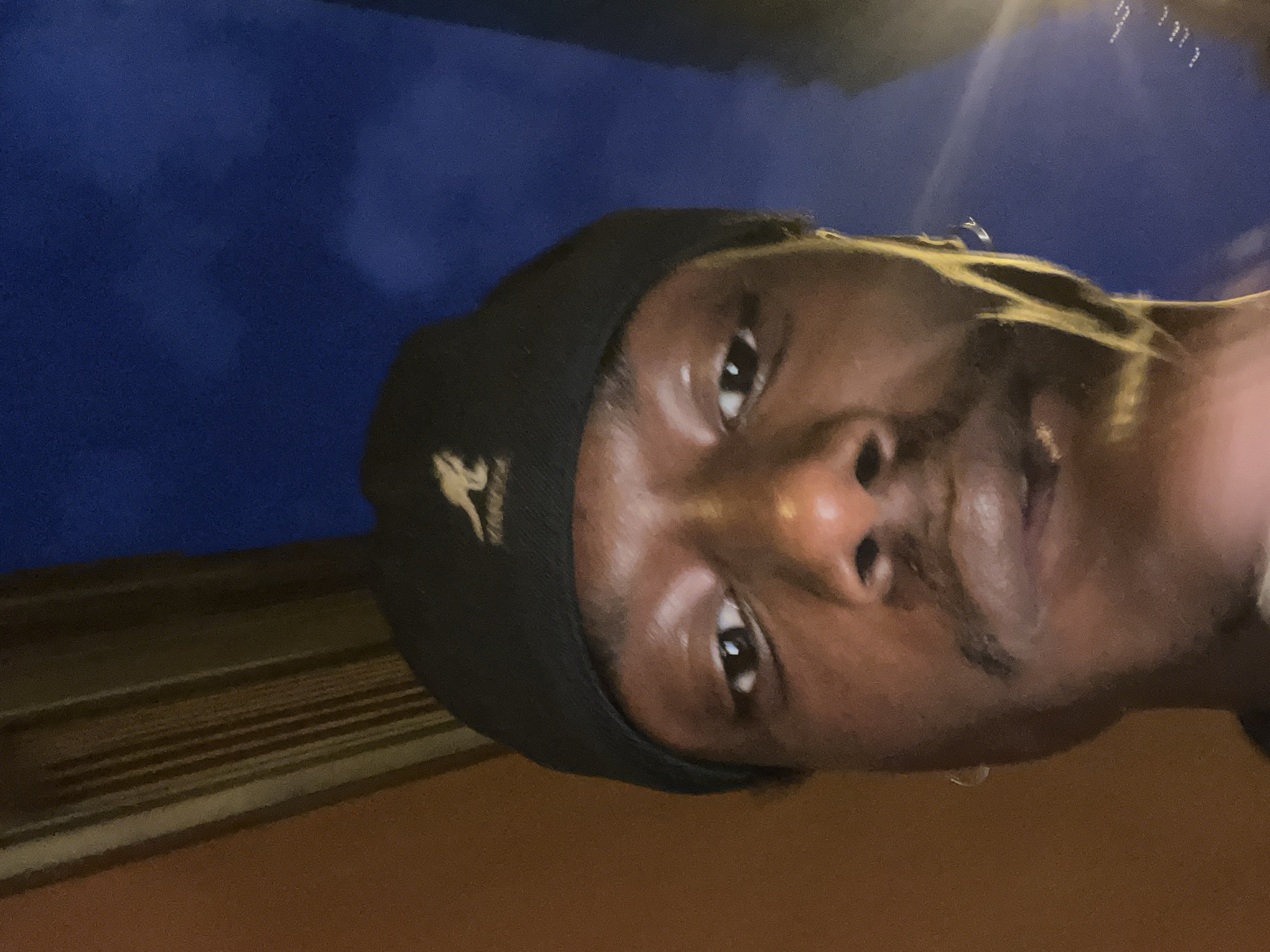 A close-up, slightly angled upward, shows a person with dark skin wearing a black beanie with a white kangaroo logo. The person's face is central, with their eyes looking directly at the viewer. The background is divided into a blue area at the top left and a brown area at the bottom. A wooden texture is visible in the brown area, running horizontally. The image is slightly blurry.
There are no elements to suggest that this image was taken in the city of Barcelona, Spain.