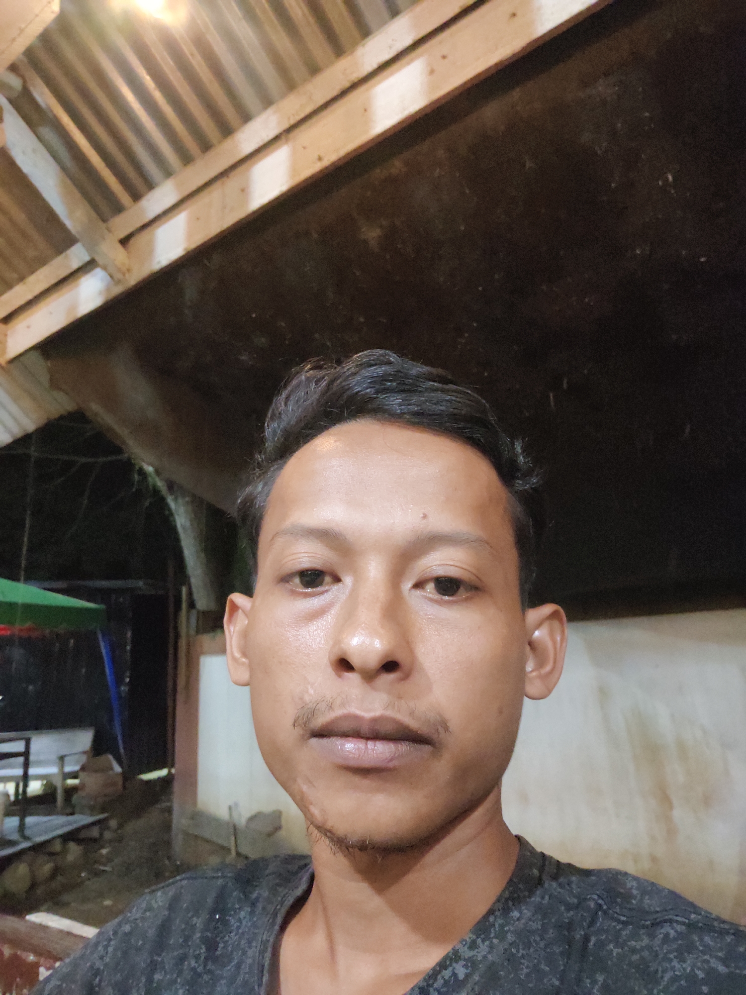 A man is taking a selfie outdoors at night. He is looking directly at the camera with a neutral expression. He has short, dark hair and a light mustache and stubble. He is wearing a dark grey t-shirt. Behind him, there is a corrugated metal roof supported by wooden beams. The background is dark, suggesting it is nighttime, and includes some out-of-focus elements such as trees and what appears to be a tent or canopy. The setting seems to be a rustic outdoor area, possibly in a rural location. There are no visible signs of text. The provided location context is Keudematangkuli, Indonesia.