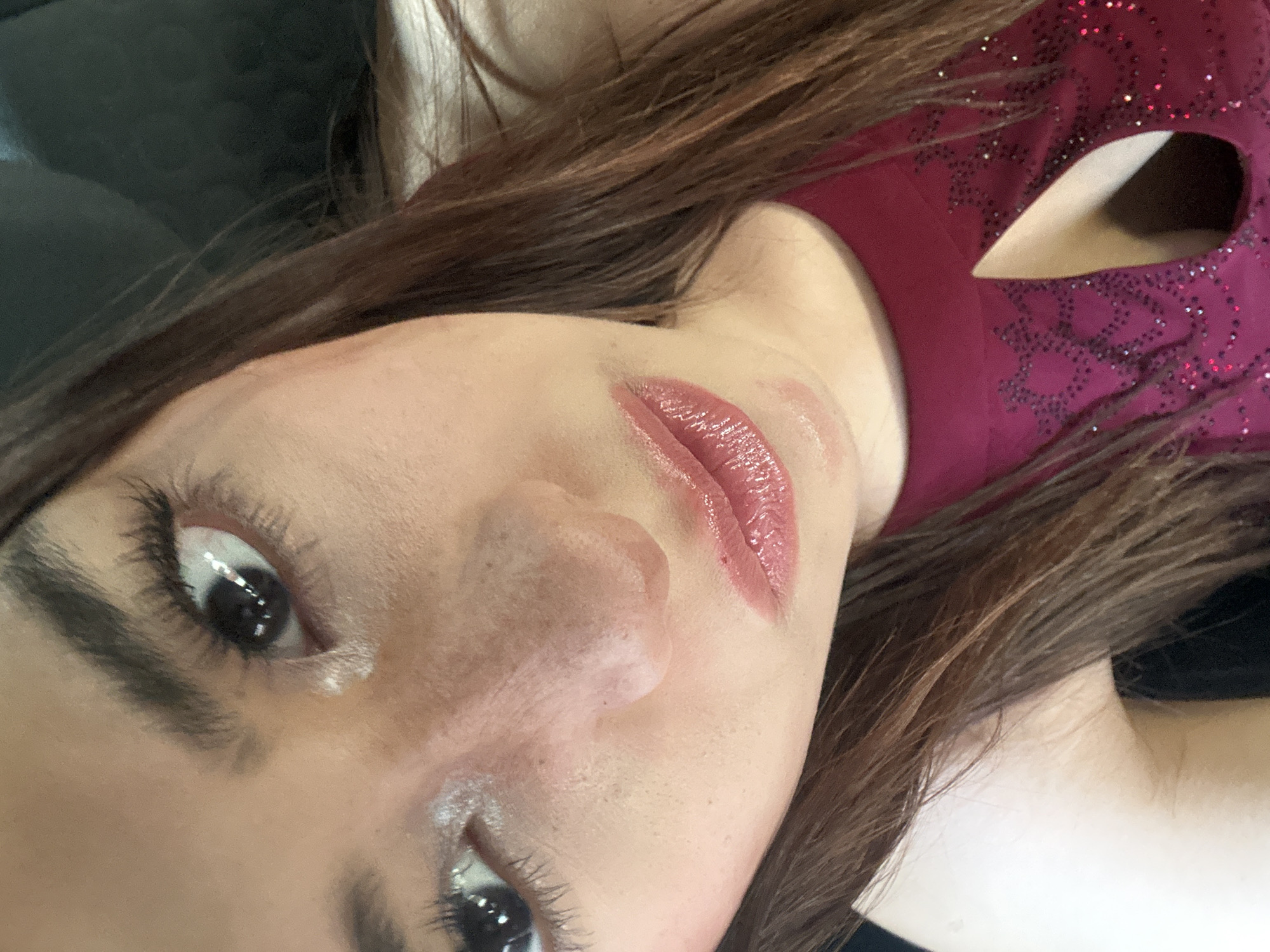 The image is a close-up, angled shot of a person's face, likely female, lying down. She has brown hair and fair skin. She is wearing a red top with a jeweled pattern and a teardrop cutout. Her lips are glossy with lipstick, and there is some smudging of the lipstick on her face. The background appears to be a dark, possibly black, surface. There is no information in the image to confirm a location in Puebla, Mexico.