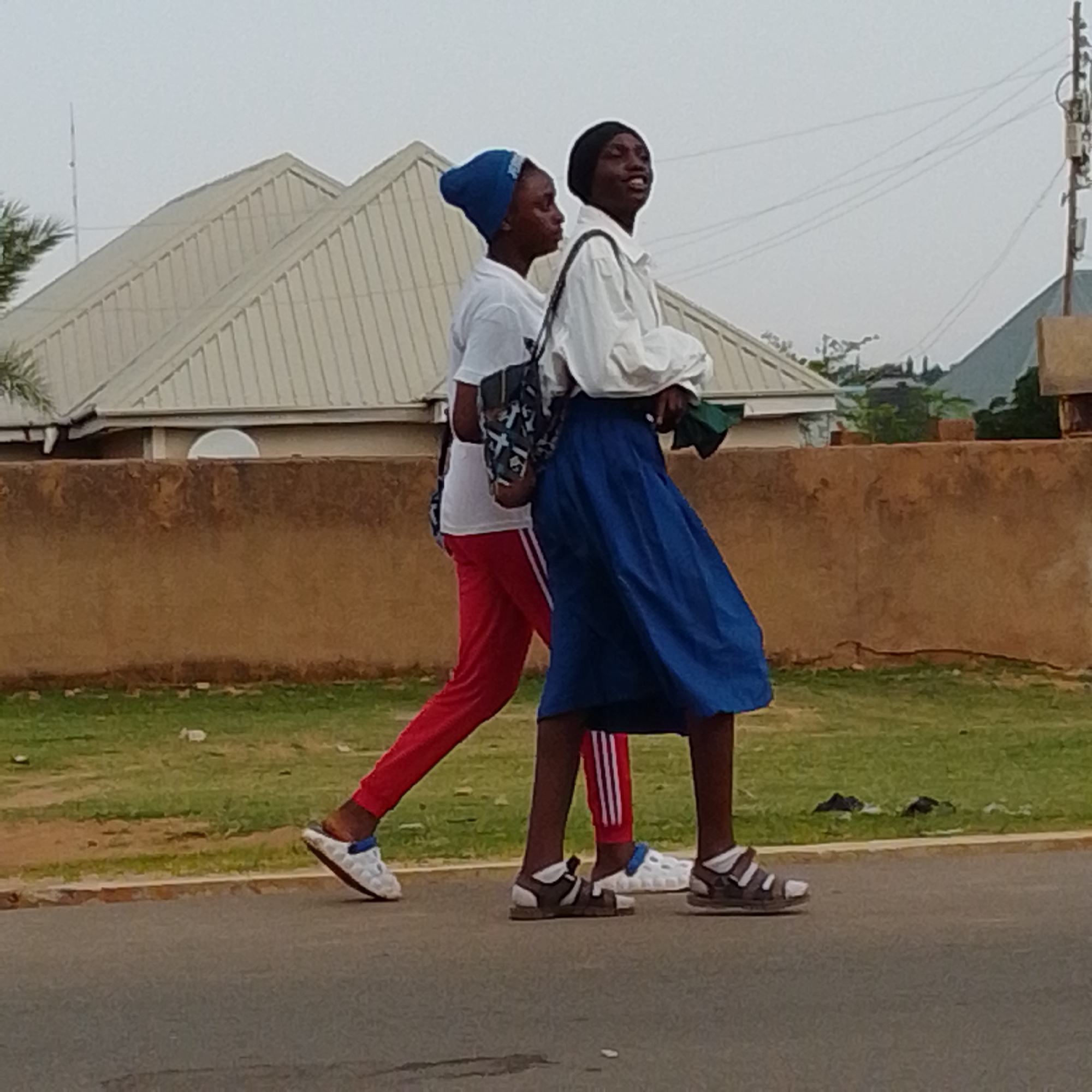 Two young people, likely teenagers, are walking on a paved road in what appears to be a residential area in Gombe, Nigeria. The person on the left is wearing a white t-shirt, red pants with white stripes, and white shoes with blue accents. They are also wearing a blue head covering. The person on the right is wearing a white long-sleeved shirt, a blue skirt, and dark sandals with white accents. They have a dark head covering. Both individuals are carrying bags. The person on the left has a patterned bag slung over their shoulder, and the person on the right is holding a green item in their hand, possibly another bag or a piece of cloth.
Behind the individuals, a low, brown-colored wall runs horizontally across the image, with a strip of green grass and some scattered debris in front of it. Beyond the wall, there is a building with a silver, corrugated metal roof, featuring a prominent gable. The sky is overcast, suggesting cloudy or possibly hazy weather. The overall lighting indicates it could be daytime, but the lack of direct sunlight and clear shadows suggests it might be an early morning or late afternoon. The presence of buildings with distinct roofing styles and the type of wall are common visual cues in Nigerian environments. The scene is a candid snapshot of everyday life, with no explicit text visible in the image. The expressions on their faces are difficult to discern clearly, but the person on the right appears to be looking upwards and to the side.
