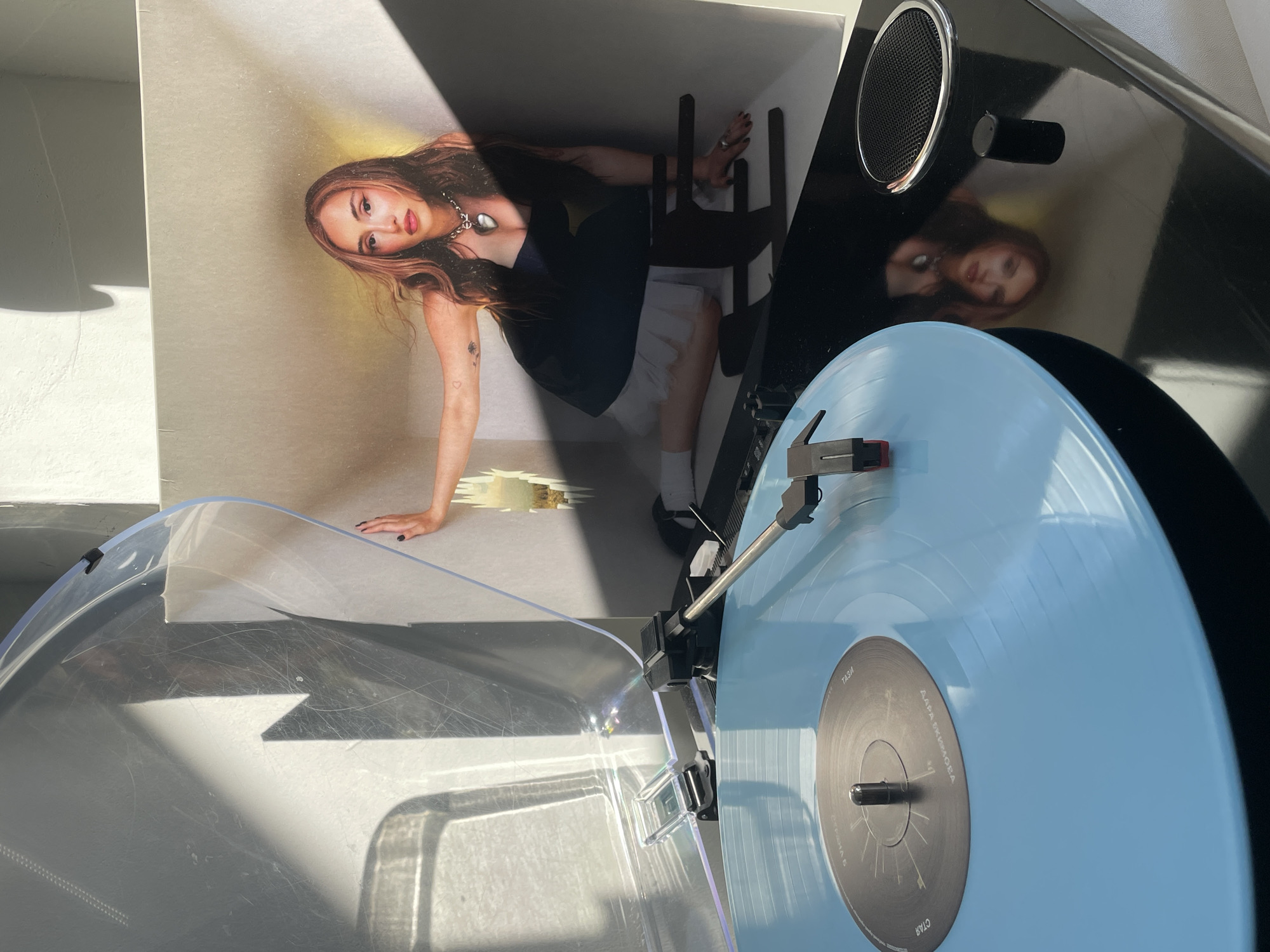 A turntable plays a blue vinyl record with a black center. The album cover sits beside the turntable and features a woman in a dark top and white skirt. She is resting on her hand against the floor. The scene is indoors with sunlight streaming in.