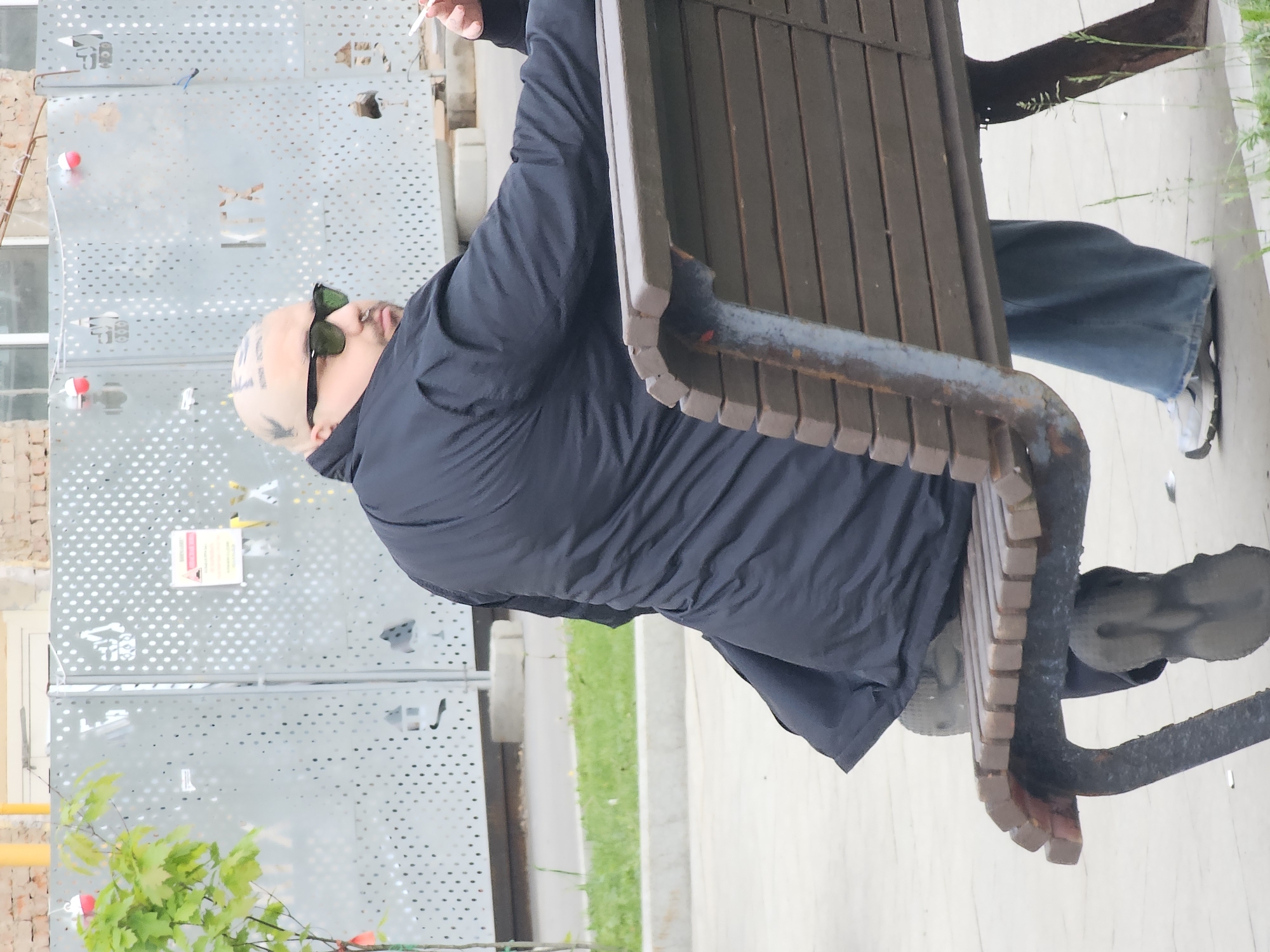 A bald man with face and head tattoos wearing dark sunglasses and a dark coat is sitting on a wooden bench outdoors in Khimki, Russia. He is smoking and his legs are stretched out. The background includes a building with a perforated metal facade, green foliage, and a sidewalk.