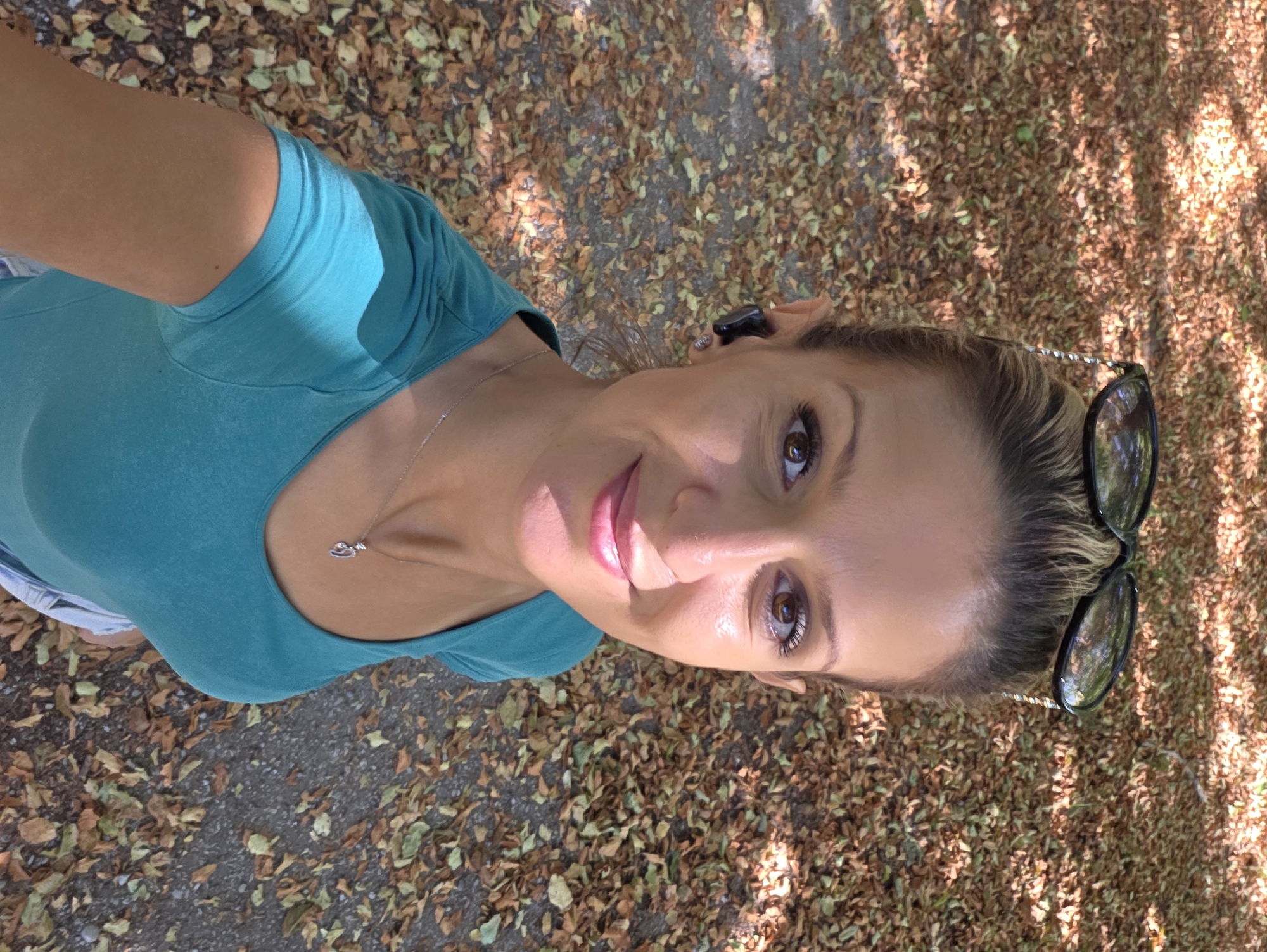 A woman is taking a selfie from a high angle outdoors. She is wearing a teal t-shirt and a necklace with a heart-shaped pendant. Her hair is pulled back and she is wearing dark-rimmed sunglasses on top of her head. She has earbuds in her ears. The background is a path covered in fallen leaves. The lighting suggests it is daytime with sunlight dappling through trees. There is no visible indication of the location being Munich, Germany.