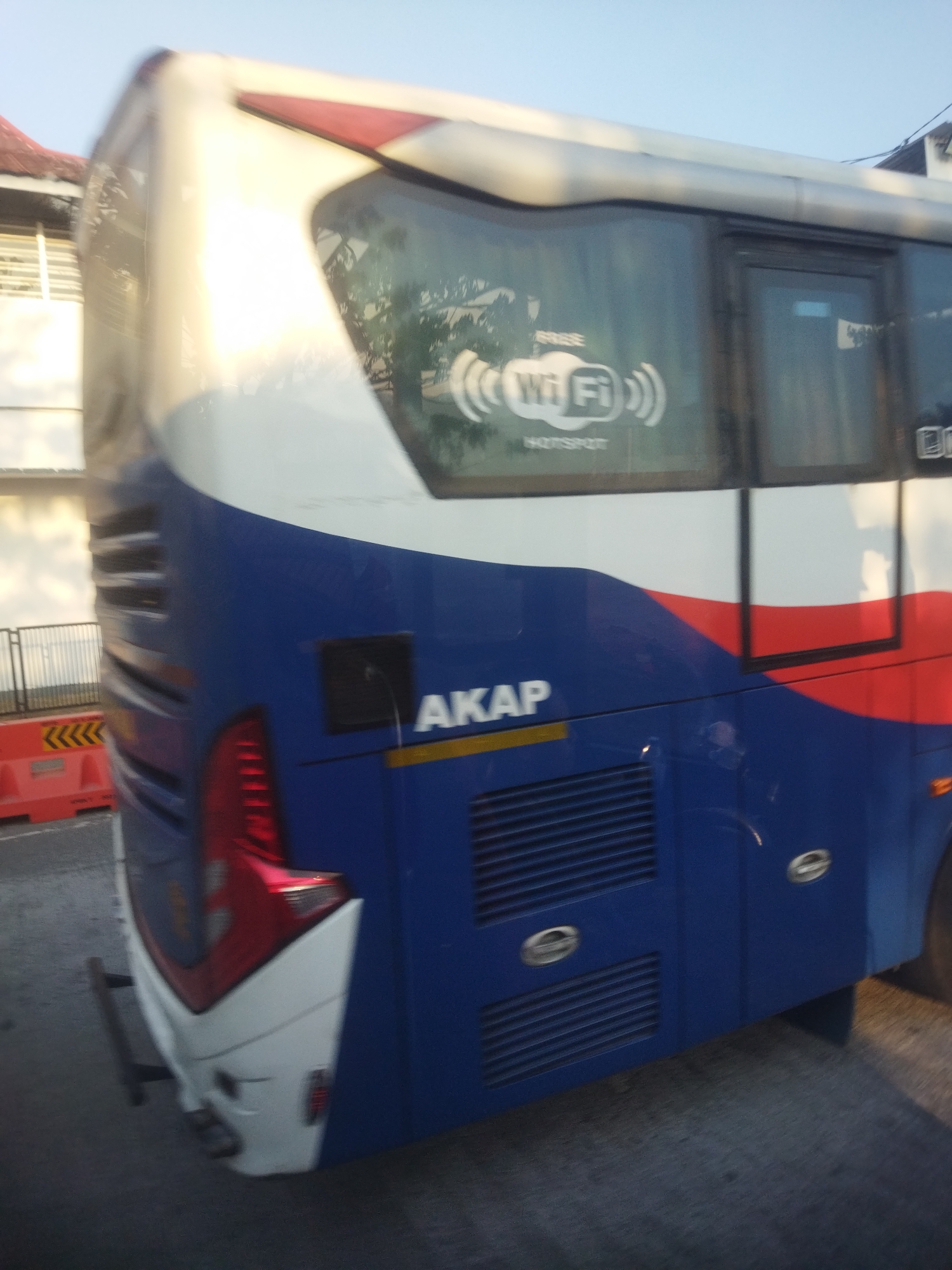 The image shows the side of a blue and white bus in Kaliancar, Indonesia. The bus has a red stripe along the top. On the side of the bus, there is a white sticker with "Wi Fi" and a hotspot symbol. The bus also has "AKAP" written in white letters. There are orange traffic barriers and a metal fence in the background. The sky is clear and blue. The image is taken from a slight angle, showing the rear portion of the bus.