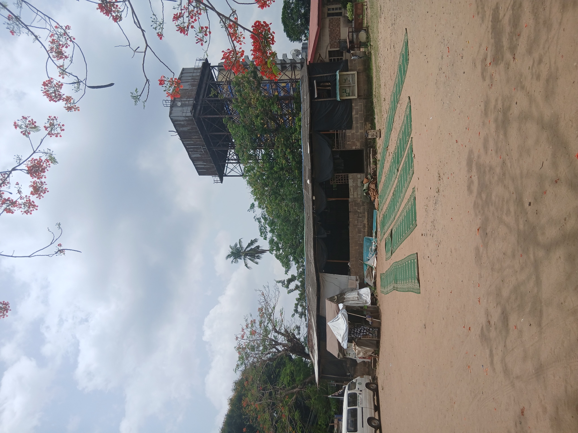The image presents an outdoor scene in Lagos, Nigeria, capturing a sandy area adjacent to a building with a distinctive water tower structure. The building appears to be constructed of concrete or brick with a corrugated metal roof. Several green patterned mats are laid out on the sand. A white van is parked next to the building, partially obscured by trees. The sky is overcast. The presence of a water tower and the building’s architecture is typical of Lagos’s infrastructure.