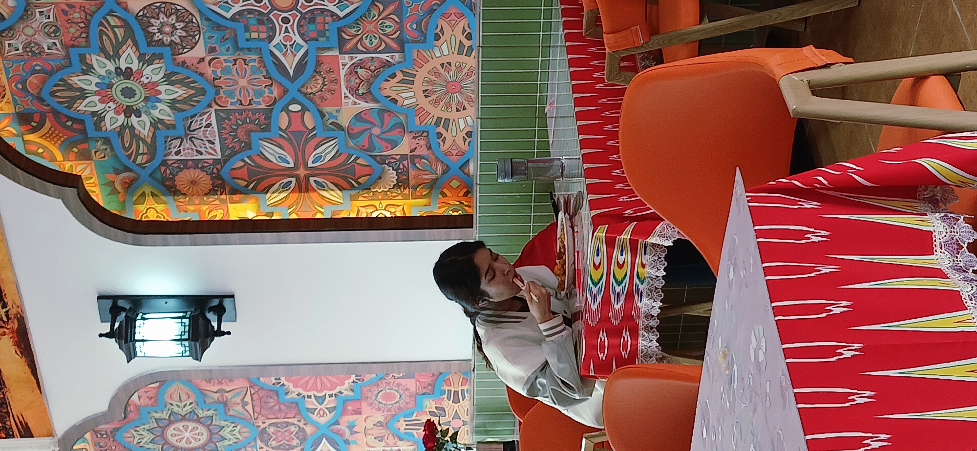 The image shows a woman sitting at a table in a restaurant, possibly in the Chongqing High-tech Zone, China. She is eating with chopsticks, wearing a white and gray jacket. The table has a red and white patterned tablecloth with lace trim. The chairs are orange. Behind her, the walls are decorated with colorful tile patterns and there's a decorative ceiling light fixture. A green wall panel is visible to the side.
