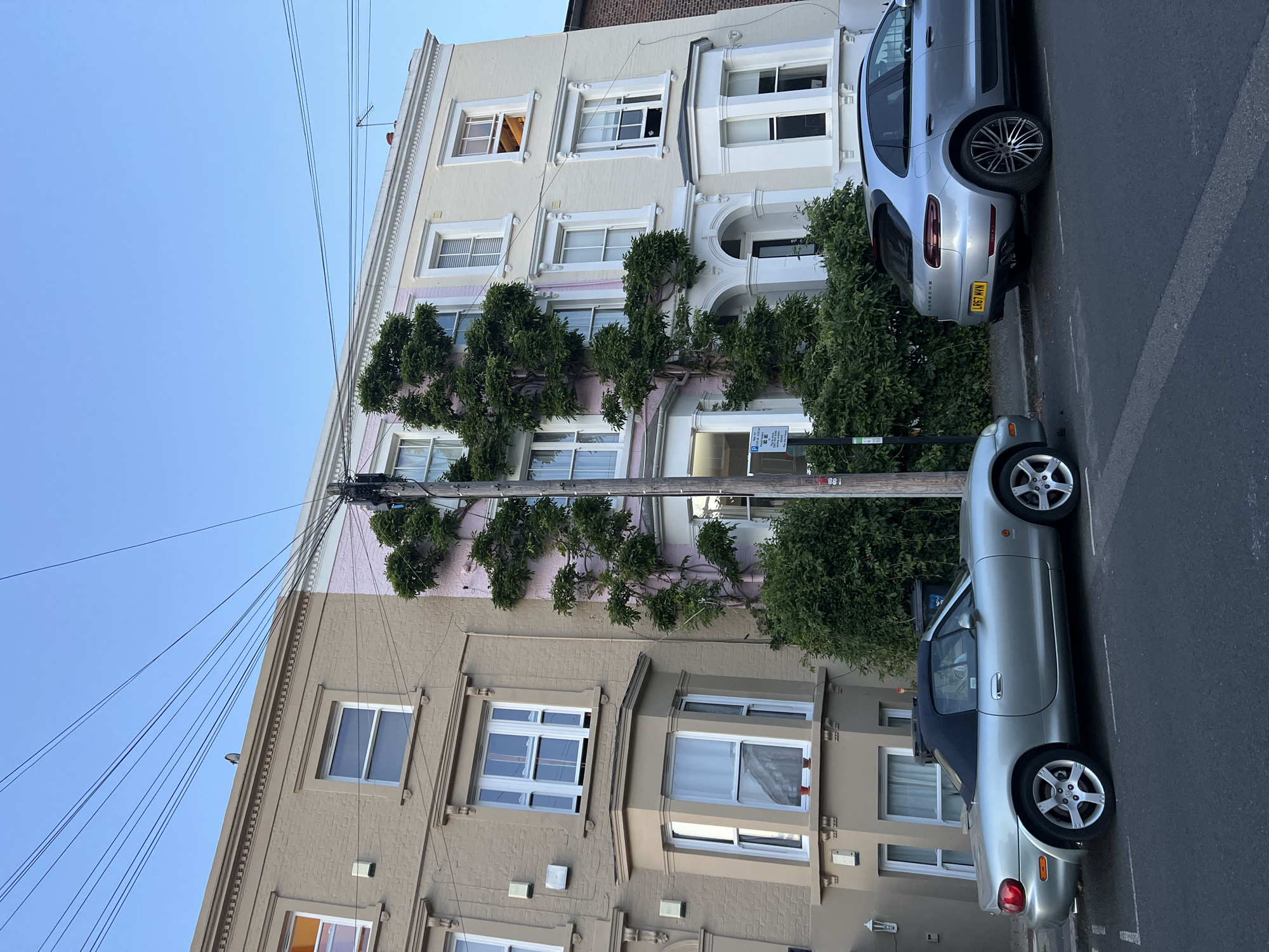 The image depicts a street scene in London, United Kingdom, featuring two parked cars and a residential building. The building is a multi-story structure with distinct architectural details, including framed windows and decorative moldings. A climbing plant, possibly ivy, adorns the facade between the building's levels. Power lines and a utility pole are visible, intersecting the building's edge. The cars parked along the street are a silver convertible and a silver Porsche SUV. The sky is clear and blue.