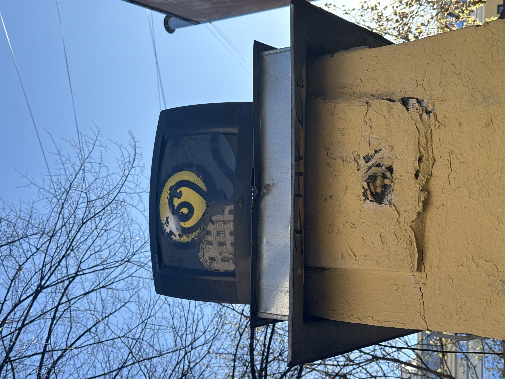 An upward-angled, low-shot photograph captures an urban scene under a clear, bright blue sky in Saint Petersburg, Russia.
The primary subjects are a large, weathered, yellow-painted wall or pillar on the right, and a dark, monitor-like object on the left. The yellow structure shows significant signs of age and wear, with cracked, peeling paint and areas where the underlying material is exposed, revealing dark patches and damage. Its surface is heavily textured and uneven.
Attached to this yellow structure by a metallic bracket is a black object resembling an old display screen or television monitor. Its surface is adorned with bold, yellow graffiti, forming a stylized symbol that appears to be a number "6" with an accompanying flourish. Above this graffiti, a faint "LG" logo is discernible on the monitor's frame. The reflective screen of the monitor clearly mirrors a light-colored, multi-story building with multiple windows, providing a glimpse of the surrounding architecture.
Connecting the yellow pillar and the black display is a silver metallic support. Faint, vertical, indecipherable text is present on the side of this silver bracket. In the background, against the vibrant blue sky, are numerous bare tree branches, indicating a colder season such as late autumn, winter, or early spring. Several thin electrical wires are also visible stretching across the sky. The overall scene presents a static, urban environment characterized by aged infrastructure and clear daylight.