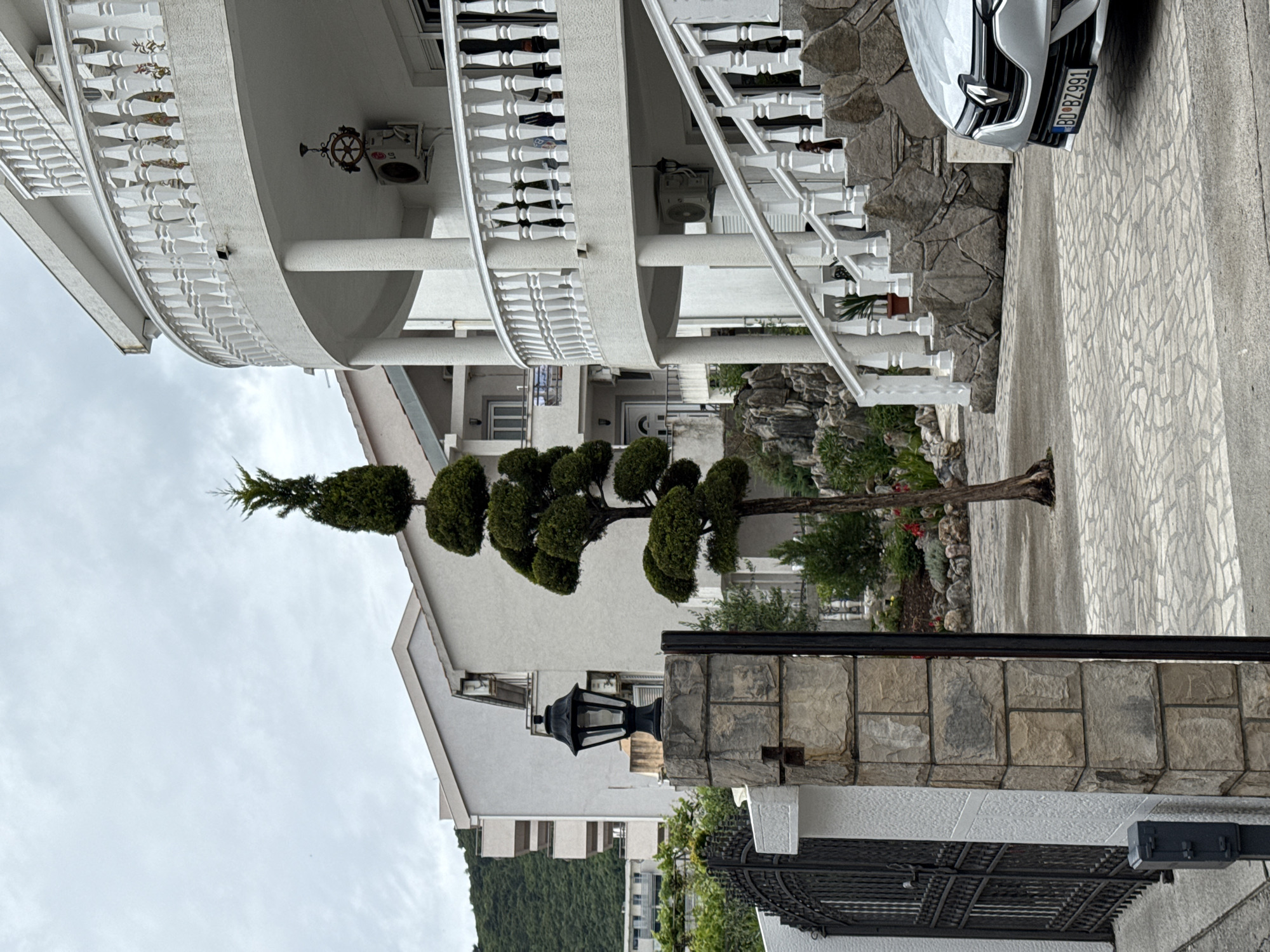 A landscape-oriented photograph shows a residential scene in Budva, Montenegro, featuring a multi-story white building with curved balconies and a decorative ship's wheel. The building has white railings and columns. To the right of the building is a paved driveway with a white car parked. The car's license plate reads "BD BZ991". A uniquely shaped tree with rounded, sculpted sections is prominently displayed in front of the building. A stone wall with a black metal gate runs along the bottom of the frame, with a black lamppost situated on top of the wall. Further in the background, a hillside covered with green vegetation is visible. The sky is overcast, suggesting a cloudy or dull day.