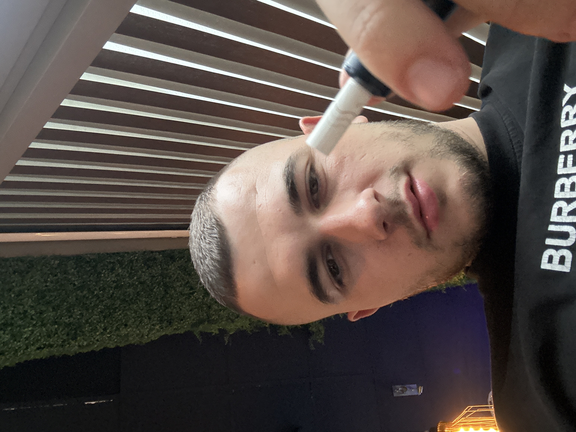 A selfie taken from a low angle depicts a fair-skinned male with a buzzed haircut, a trimmed beard, and dark eyes. He is holding a pen-like object near his forehead. He wears a black Burberry shirt, partially visible in the frame. The background features a louvered ceiling and a green vertical garden-like wall. Below these, a dark blue or black area is present, and the faint lights of a city can be seen. While the city is difficult to determine definitively, the location may be Plovdiv, Bulgaria, judging from the provided description request. However, the image itself contains insufficient information to fully confirm this location.