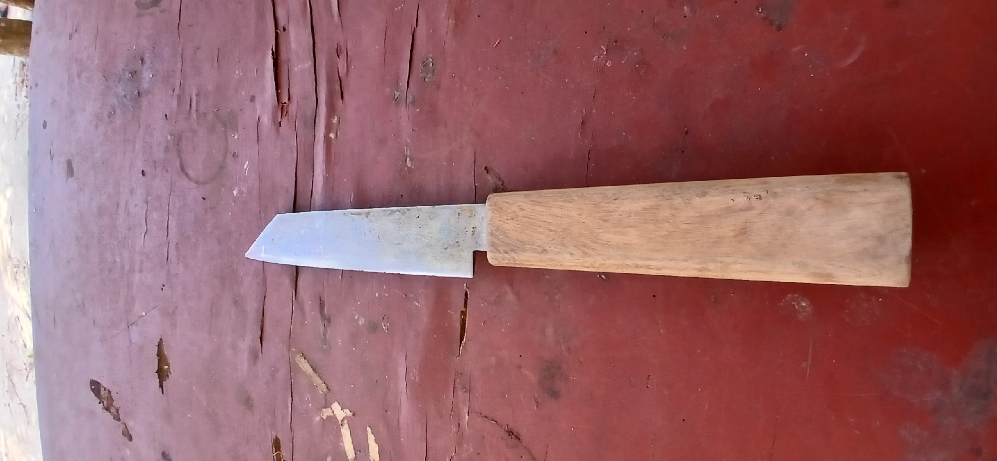 A knife with a metal blade and a wooden handle rests on a weathered, red wooden surface. The blade is angled at the tip, and the wooden handle is rectangular with slightly rounded edges. The surface beneath the knife is textured with cracks and peeling paint, suggesting it is an outdoor or well-used item. The lighting indicates it is daytime, with a slight shadow cast by the knife. The location is described as Pejaten, Indonesia.