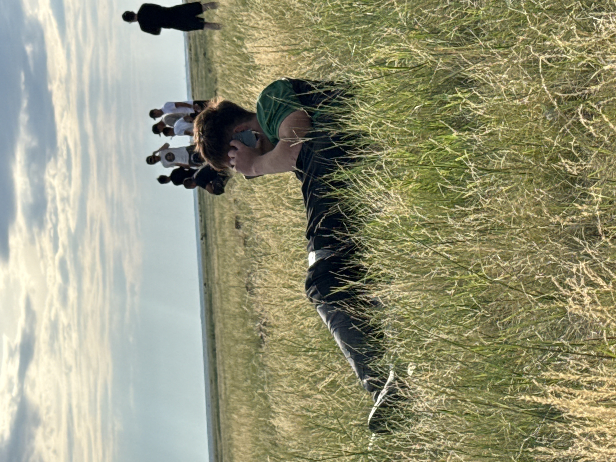 The image depicts a man lying in tall grass, holding a cell phone to his ear. He is wearing a green shirt and dark pants. In the background, a group of people are standing and looking in the distance. The environment appears to be a vast, grassy plain under a cloudy sky. The location is identified as the city of Өндіріс, Kazakhstan. The scene is open and rural, with no visible urban structures. The lighting suggests it is daytime, likely late afternoon or early evening due to the soft, diffused light.