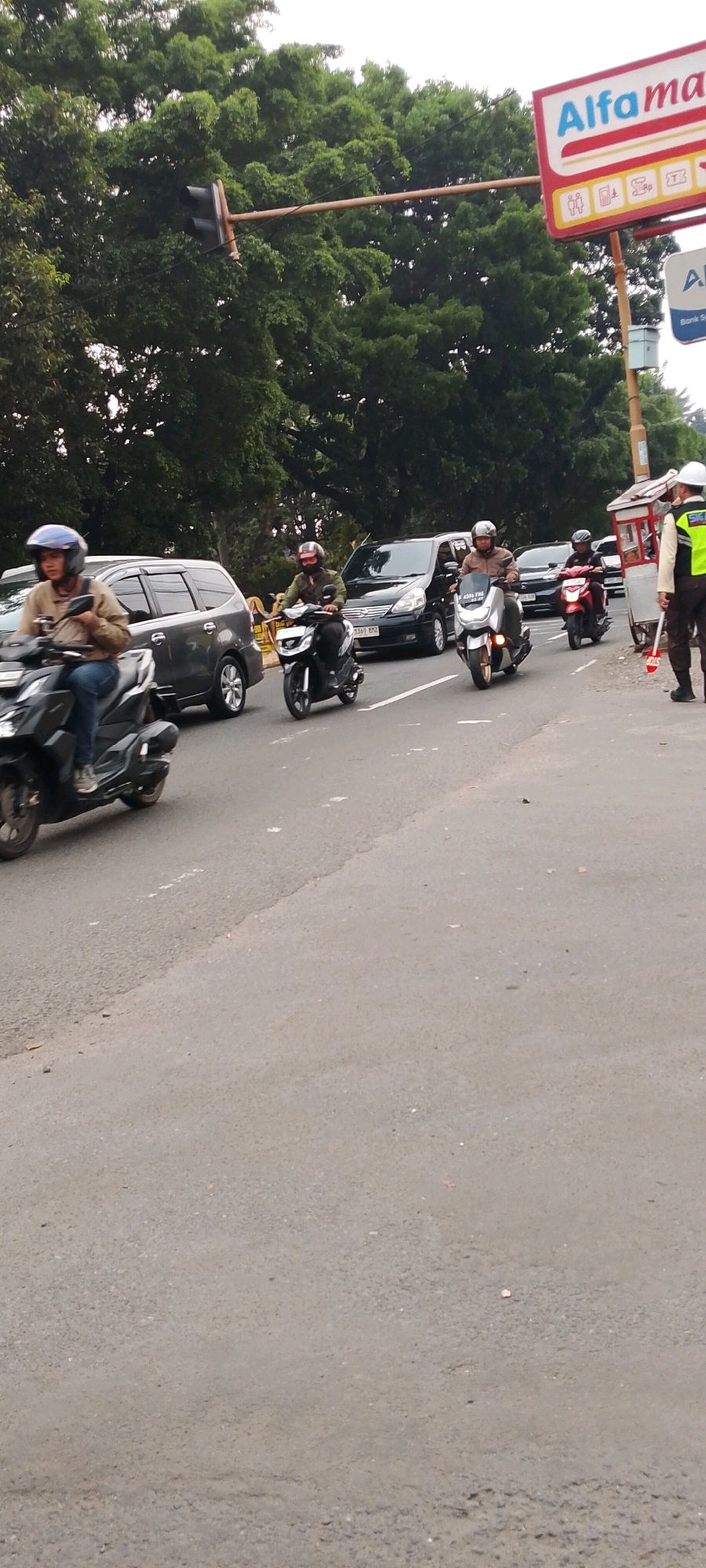 The image depicts a street scene in Depok, Indonesia, likely during daytime given the lighting conditions. Several motorbikes and cars are present, indicating moderate traffic. On the left side, a person wearing a light brown jacket and blue jeans rides a black motorbike, with a silver helmet. A dark gray minivan is to their right. In the center, another person in a green jacket and helmet rides a black motorbike. Behind them, a silver motorbike is being ridden by someone in a brown jacket and helmet. Further back, a black car is visible. To the right of the road, a person in a high-visibility yellow and black vest, likely a traffic officer, stands near a red and white cart. A red motorbike is also seen in this area. Overhead, a traffic light is visible, and a sign for "Alfamart" is prominently displayed on the right. The environment includes lush green trees in the background, suggesting a location with ample vegetation. The road surface is asphalt.