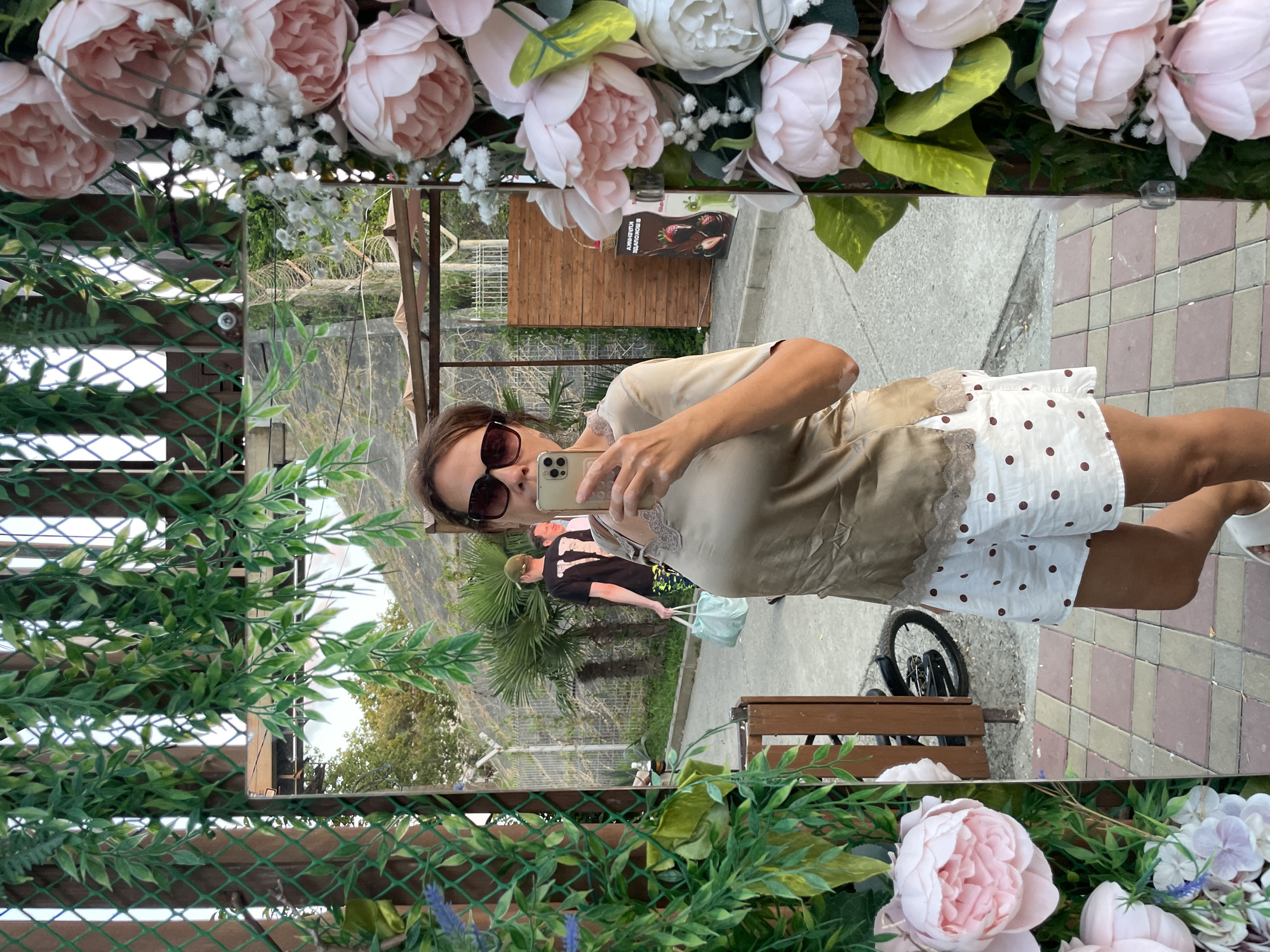 A woman in her 40s, wearing sunglasses, a light brown silk blouse with lace trim, and white polka-dot shorts, stands and takes a selfie in a tall, ornate mirror. She is holding a smartphone in her right hand. The mirror is framed by artificial greenery and pink and white flowers. In the reflection, a man in a black t-shirt with the number "17" on it and a green cap is walking away from the camera carrying a light blue bag. Behind him, another man is visible. The background of the reflection shows a paved walkway, a fence, palm trees, and a wooden structure. The scene appears to be outdoors, likely in a public area such as a promenade or park, possibly in Sochi, Russia, given the presence of palm trees and the style of the walkway.