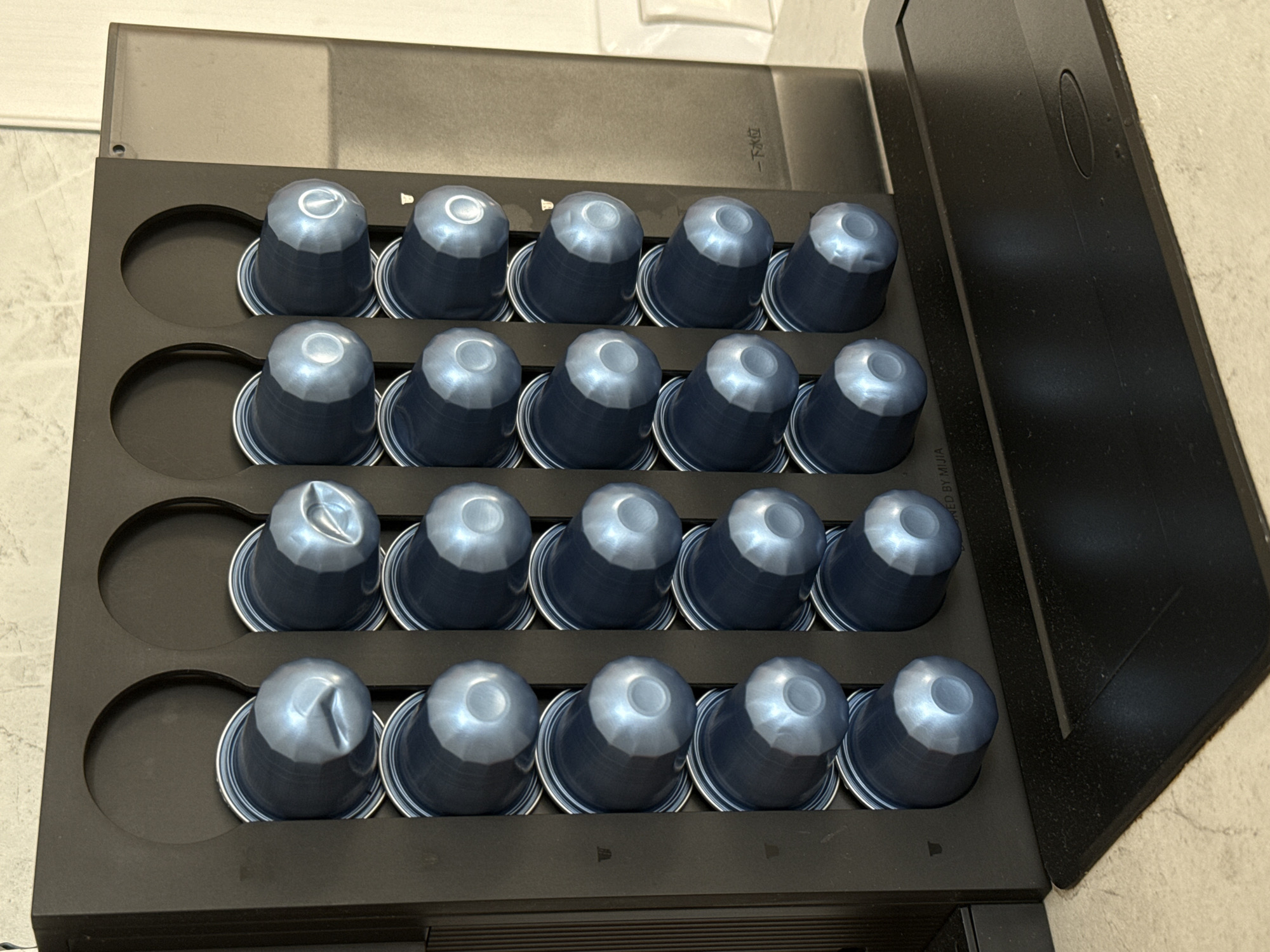 A coffee capsule dispenser is shown in an indoor setting. The dispenser is made of black plastic and has 20 slots arranged in four rows of five. Each slot holds a single blue and silver coffee capsule. The dispenser is positioned on a black tray, which is on a light-colored countertop. In the background, a white wall and a portion of a metallic appliance are visible. There is text on the right side of the metallic appliance. The image does not depict any people or indicate a specific location within the city of Lyubertsy, Russia.