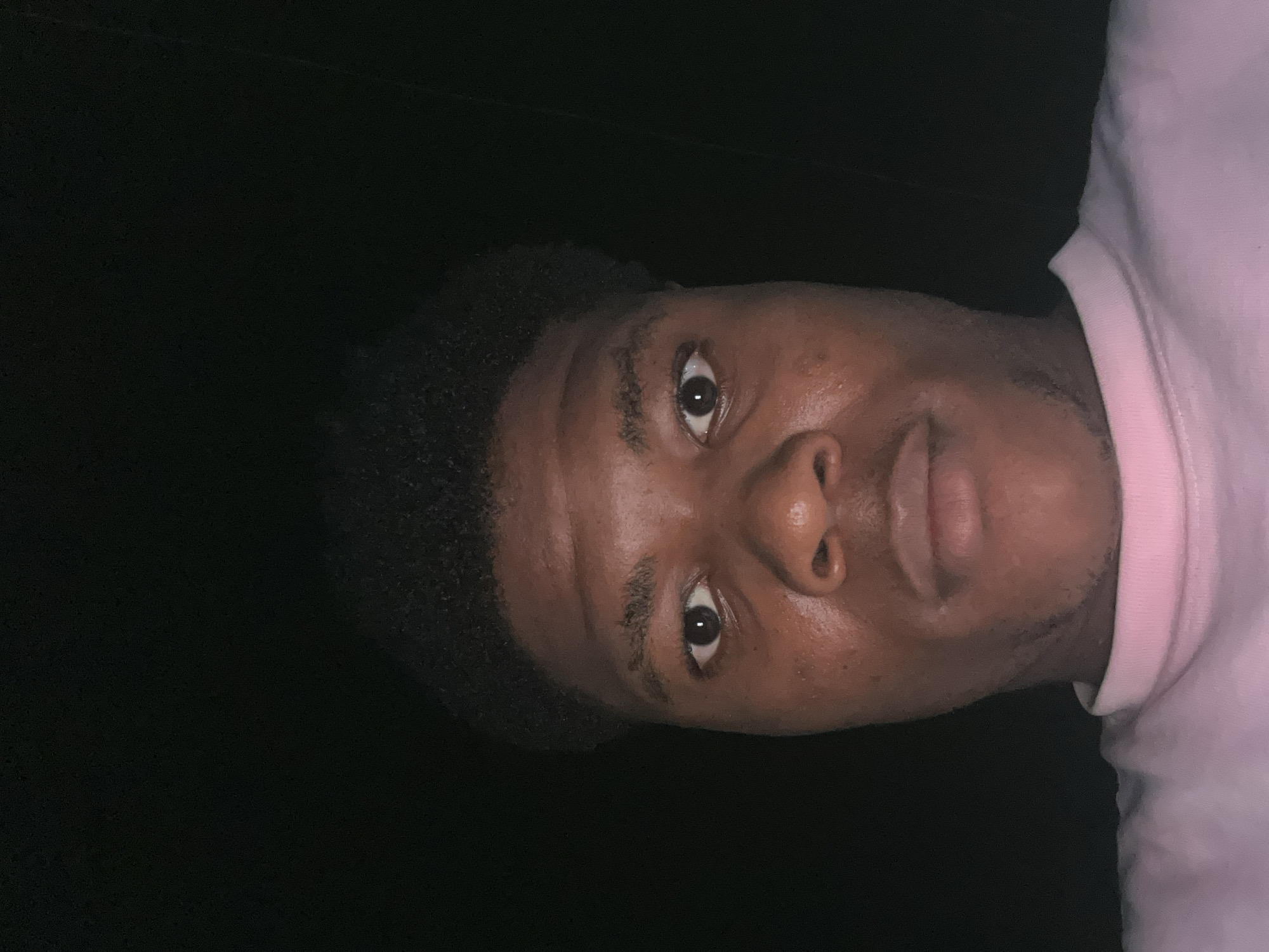 The image is a close-up, low-angle selfie of a Black male with dark, curly hair and brown eyes. He is wearing a light pink, crew-neck shirt. His skin appears to have a reddish undertone, and there are some blemishes visible on his face. The background is uniformly dark, suggesting a dimly lit or nighttime environment. The image is taken in Ihiagwa, Nigeria, though no specific location details within the city are discernible from the visual content.