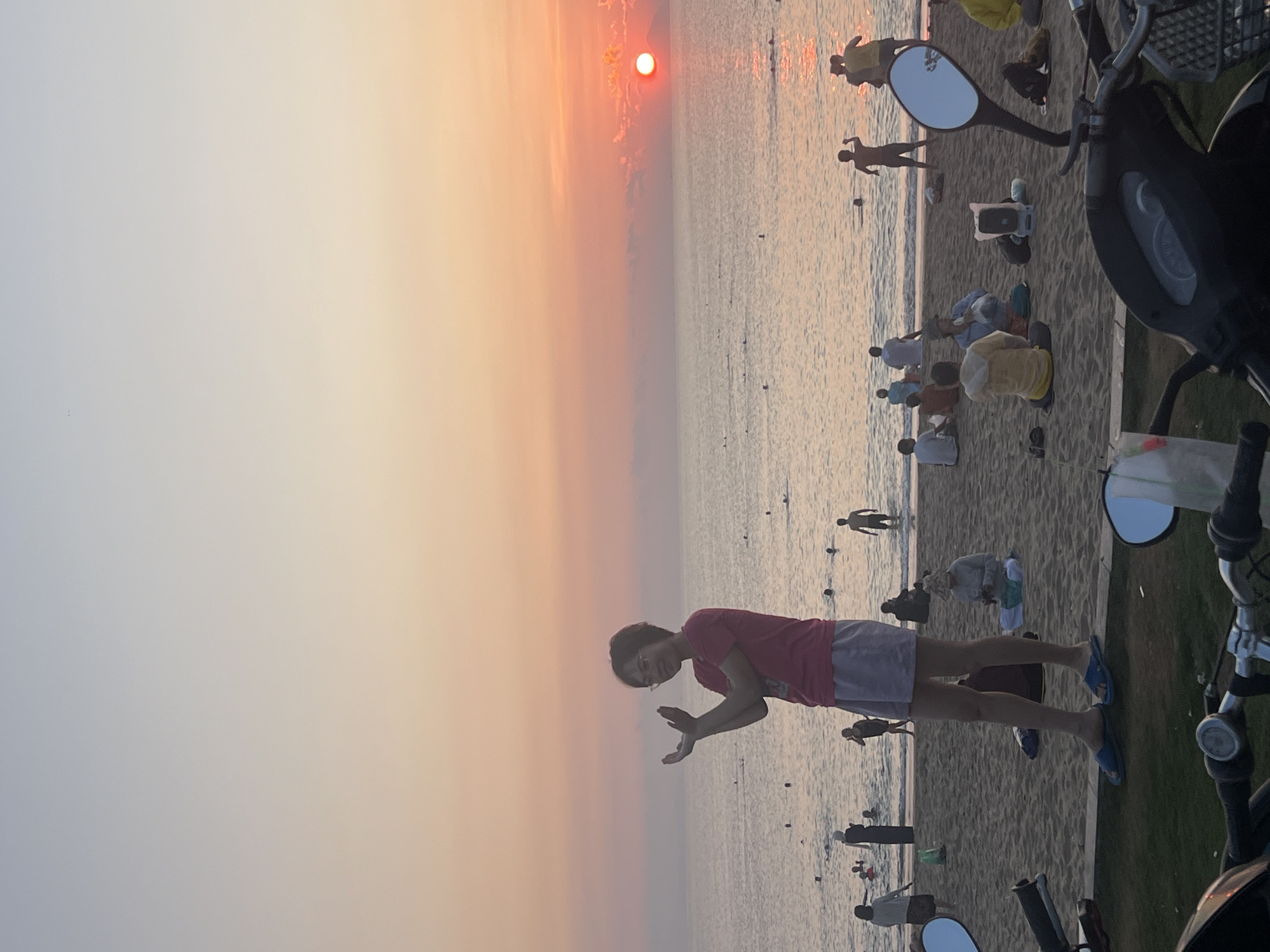 The image captures a vibrant sunset over a beach in Nha Trang, Vietnam, with the scene oriented such that the horizon runs vertically down the center. The sky on the left side is a gradient of warm colors, from a brilliant orange-red at the horizon where a distinct, glowing red sun dips into the sea, transitioning to lighter pinks and soft grey-blues higher up.
The tranquil ocean, on the right side of the horizon line, reflects the sunset's fiery hues, creating sparkling patterns on the water's surface. Numerous individuals are scattered across the sandy beach and in the shallow waters, enjoying the evening. Many are silhouetted against the bright sky and water, some sitting on the sand, others standing or wading. Their activities suggest relaxation and enjoyment of the coastal environment.
In the immediate foreground, a person, likely female, stands on the beach, facing the viewer with a slight smile. She wears a red t-shirt, light grey shorts, and blue flip-flops, with glasses on her face. Her hands are raised and bent, making a playful or gesturing pose. On the far right, parts of a dark-colored scooter or motorcycle, including handlebars, a rearview mirror, and part of the dashboard, are visible, framing the scene. The overall impression is one of a peaceful, recreational evening at the beach. No visible text is present.