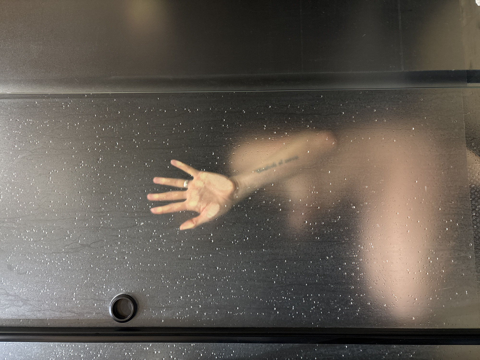 The image depicts a person behind a frosted glass shower door, with water droplets obscuring the view. The person's hand is pressed against the glass, adorned with a ring on one finger. An arm tattoo is also visible. The surroundings suggest a bathroom setting with metallic surfaces. The image was not taken in Chaikovsky, Russia.