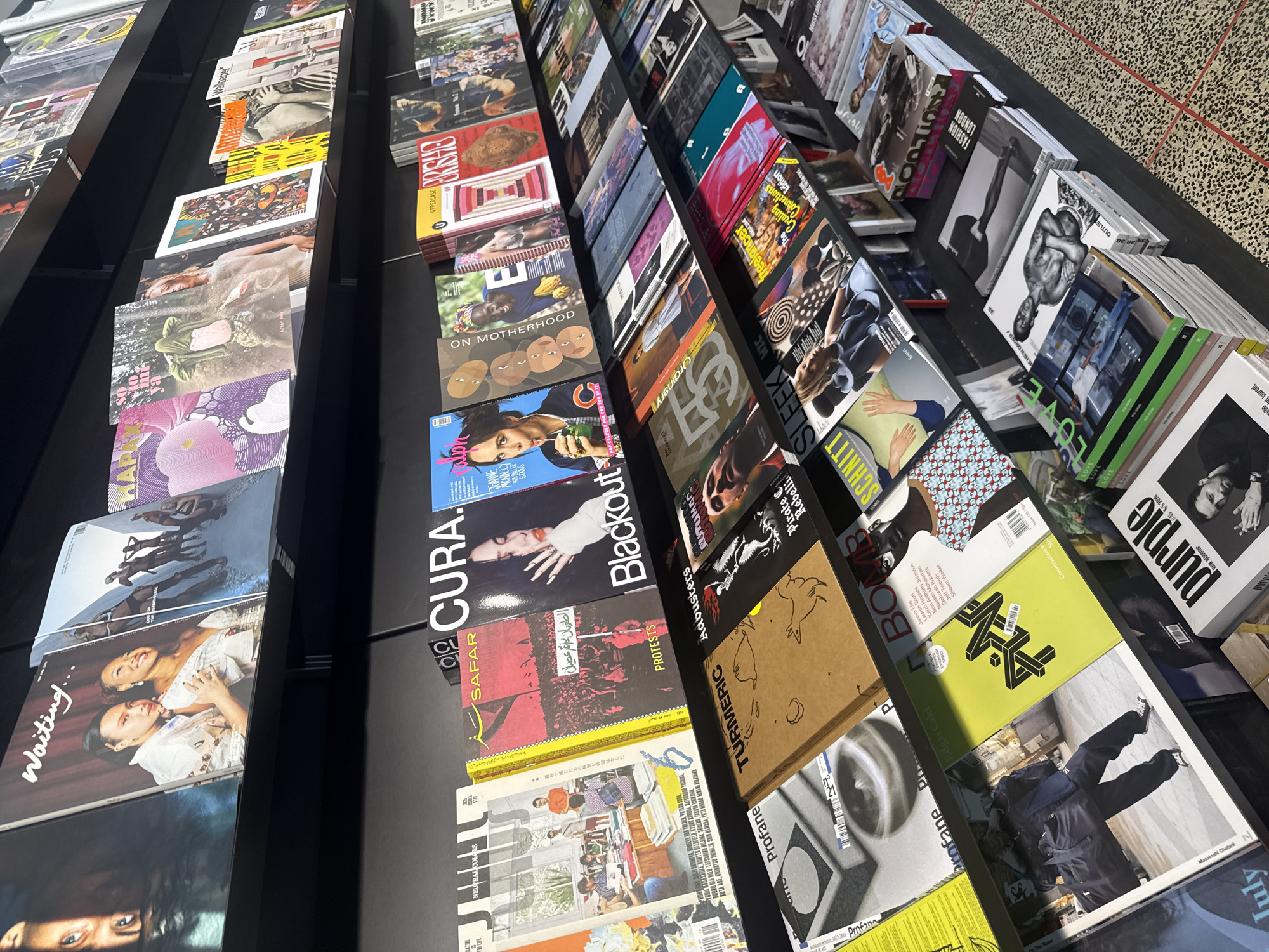 The image shows a display of magazines arranged on inclined shelves in what appears to be a bookstore or magazine shop. The magazines are organized in rows, offering a variety of titles, designs, and themes. The titles include "Waiting", "CURA", "SAFAR", "Blackout", "Protest", "Ordinary", "On Motherhood", "Adbusters", "Uppercase", "Brigitte", "Schnitt", "Sleek", "Love", "Purple", "Wallpaper", and "Fashion London." The display has a diverse aesthetic, including art, photography, fashion, and cultural topics. The location is likely in London, United Kingdom, based on the "Fashion London" magazine. The environment seems well-lit.