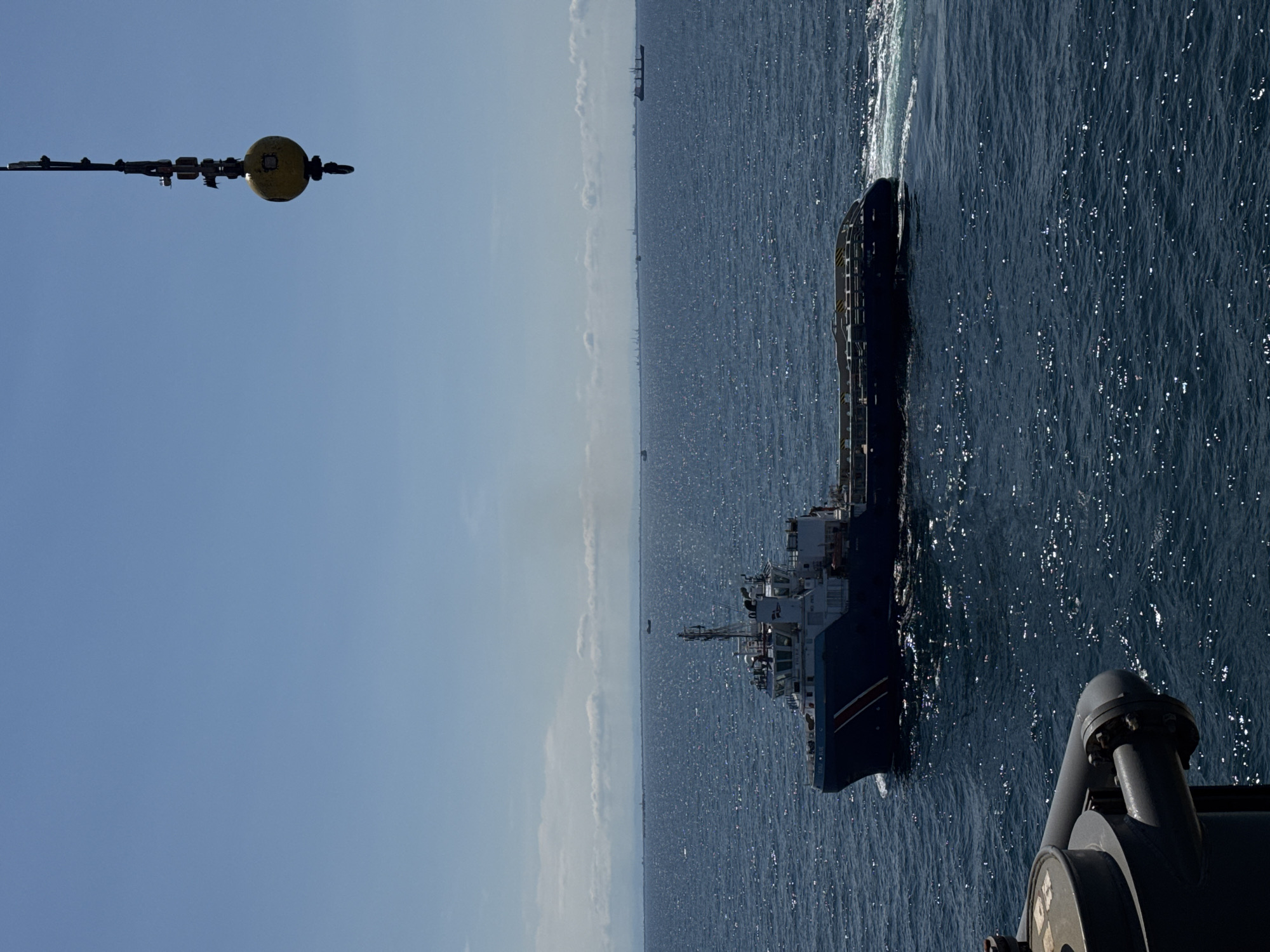The image depicts a maritime scene. In the foreground, part of a larger vessel or structure is visible, with pipes and metal components. This object is positioned on the right side of the frame. To the left of this foreground object, a yellow buoy is suspended from a boom or arm, also extending from the foreground vessel.
The main focus of the image is a medium-sized, dark blue ship moving through the water. The ship has multiple decks and appears to be a working vessel, possibly a tugboat or a supply ship, with visible structures, railings, and antennas. The ship is moving away from the viewer, towards the horizon. The water is a deep blue, with sunlight reflecting off the ripples, creating a shimmering effect.
In the distance, the horizon is visible, with a hazy, lighter blue sky. There are some light clouds scattered near the horizon. On the far left of the horizon, a very faint silhouette of what appears to be a distant coastline or a larger ship can be seen. The overall impression is a bright, clear day at sea. The time of day appears to be daytime, judging by the sunlight and the clear sky. The scene is one of calm movement on the open water, with the two vessels engaged in some form of maritime activity, possibly transferring supplies or personnel, or simply navigating. There is no visible text in the image.