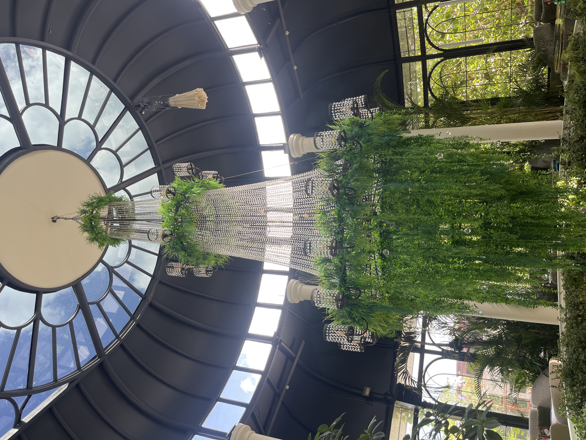 This is an interior shot taken in North Kuta, Indonesia, looking upwards towards a high, ornate ceiling. The ceiling features a large, circular glass dome, allowing a view of the sky with some clouds visible. The dome is supported by a dark metal framework with rounded edges and arched glass panels. Below the dome, a large, multi-tiered chandelier hangs, decorated with crystal strands and green foliage. The chandelier is centrally positioned within the frame. To the right of the chandelier, a series of skylights are visible in the ceiling, providing additional natural light. Tall, white columns are located on the right side of the frame, surrounded by greenery. The overall environment suggests an upscale interior, possibly a restaurant or event venue. The lighting is bright, due to the natural light sources above.