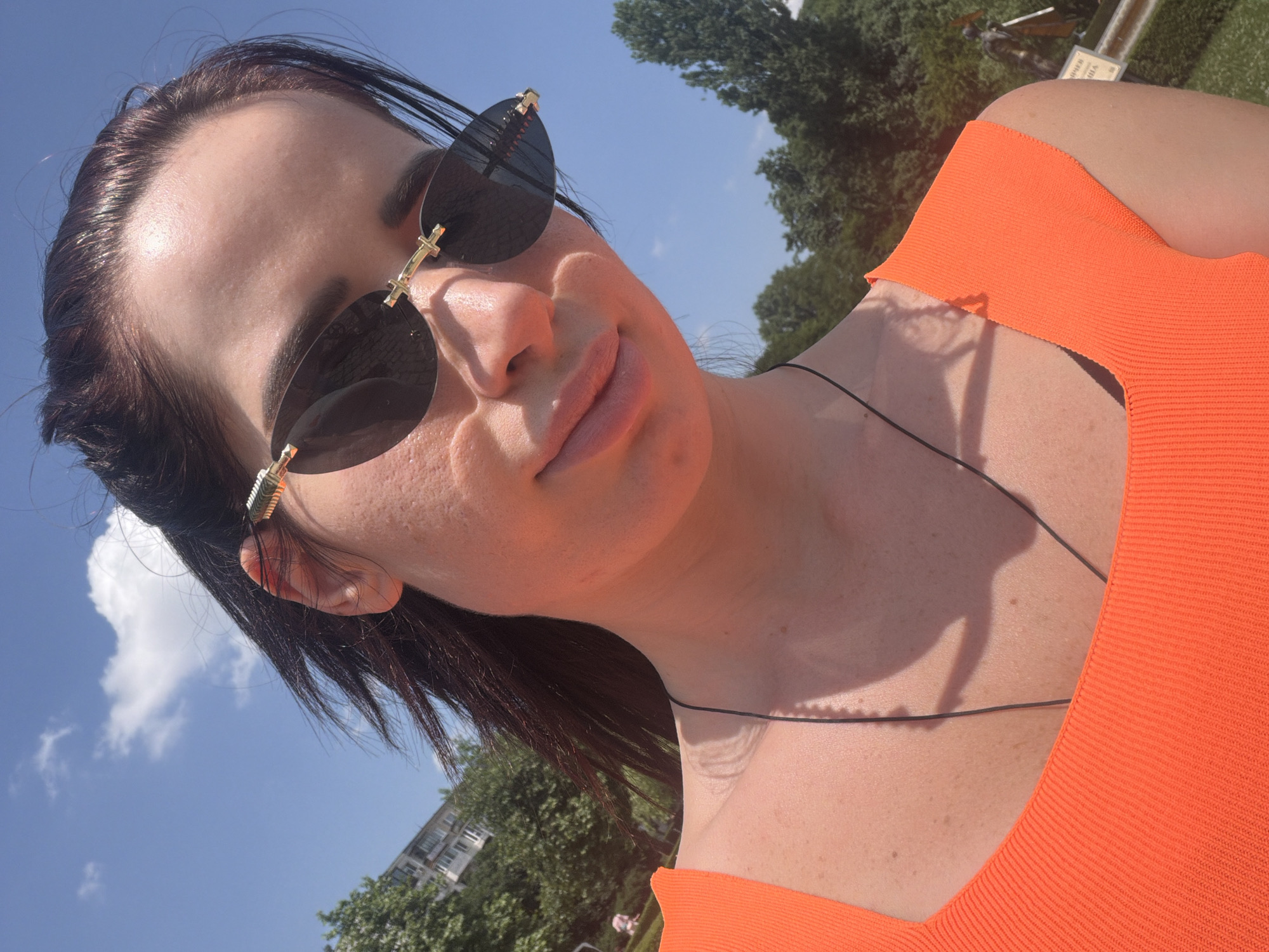 An outdoor portrait captures a woman from the chest up, looking downwards at the camera on a clear, sunny day. She wears dark, rimless sunglasses with gold-toned decorative accents at the temples and a bright orange, ribbed square-neck top. A thin black cord necklace rests on her chest. Her dark hair is styled back, revealing her forehead, which has a slight sheen, and her ears. Her skin displays visible texture on her cheeks and chin. The background consists of a vibrant blue sky with scattered white clouds, above a dense line of green trees. To the left, a portion of a light-colored building with multiple windows is visible behind the foliage. To the far right, partially obscured by trees, a faint, light-colored statue or figure stands near a rectangular sign featuring Cyrillic script, partially legible as "ВЪРНАТ" (VURNAT), indicating a location consistent with an urban park in Sofia, Bulgaria.