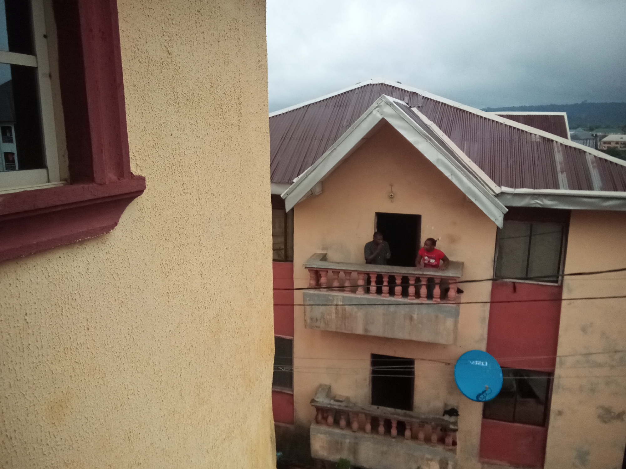 This is a medium shot, taken from a slightly elevated angle, of a multi-story building in Amaokpala, Nigeria. The building features a rust-colored corrugated metal roof with white trim. The exterior walls are a light tan color, with sections of red. There are multiple balconies with decorative railings. Two individuals are visible on a second-story balcony; one man in a dark shirt and one person wearing a red shirt. A blue satellite dish is mounted to the exterior wall of the building. In the foreground, a portion of a tan-colored wall with a dark red window frame is visible. The sky is overcast.