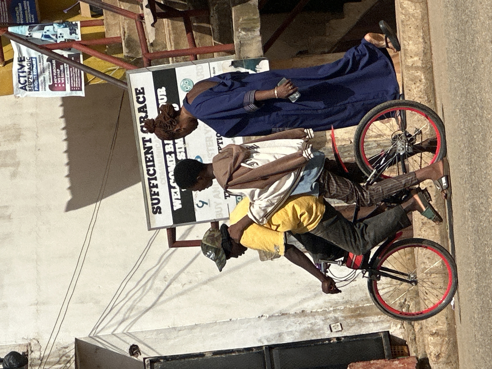 In Bukuru, Nigeria, a young boy wearing a camouflage cap and a yellow t-shirt rides a bicycle with red rims. He is carrying a white bag on his back. Behind him, a young person in a brown hooded jacket walks alongside, and a woman in a dark blue dress with an open back walks further behind, holding a phone in her right hand. A sign that reads "SUFFICIENT GRACE COMMUNICATIONS" is visible in the background, along with other signs advertising phone repairs and services. The scene appears to be on a street with a textured wall and a set of stairs.