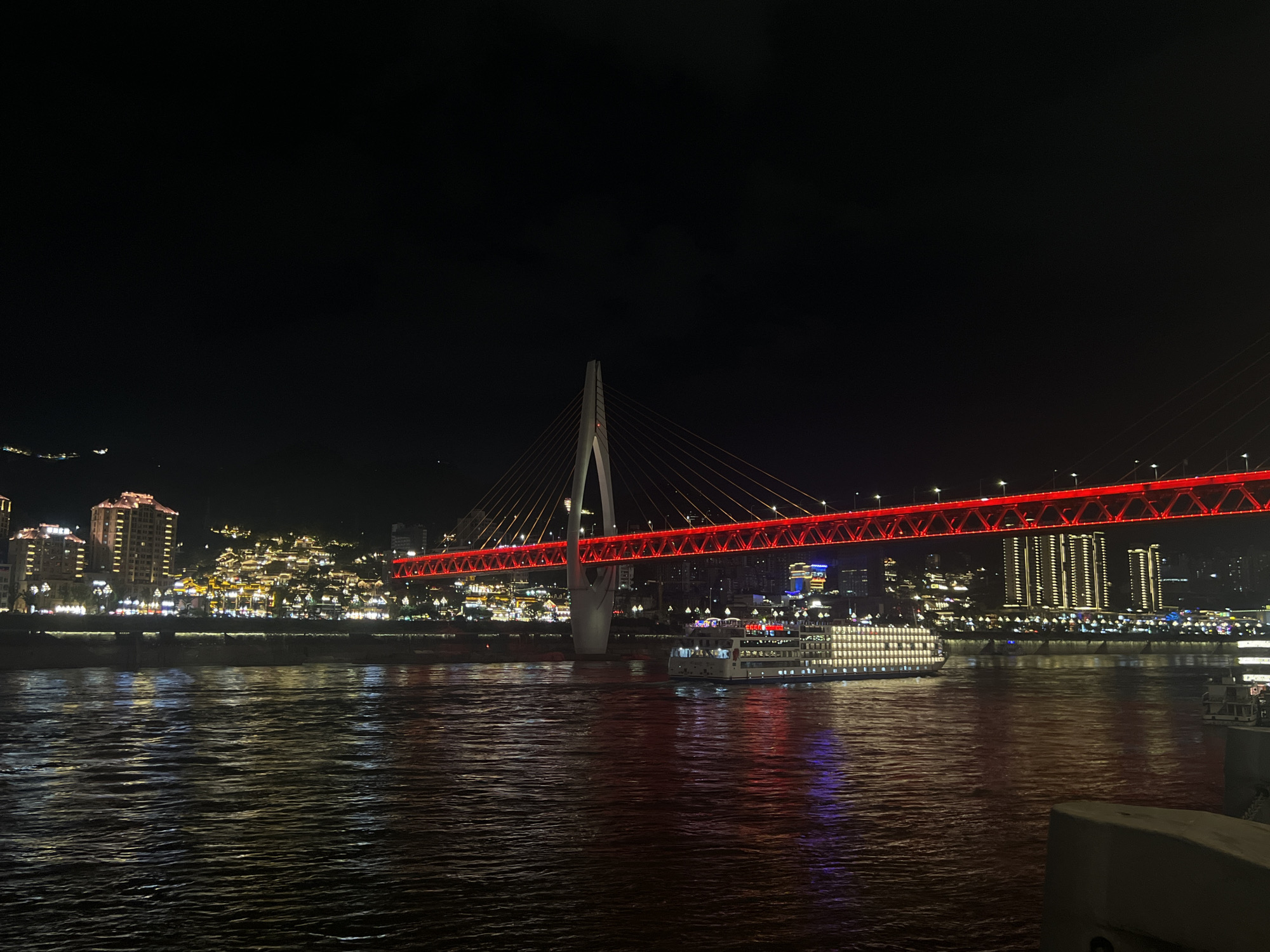 The image captures a nighttime view of a cityscape with a prominent bridge illuminated in red. The bridge is a cable-stayed bridge, with a large white pylon supporting cables that extend to the bridge deck. The bridge deck itself is lined with red lights, creating a striking visual against the dark sky.
In the foreground, a wide river reflects the lights of the city and the bridge, creating shimmering patterns on the water's surface. A large white cruise ship with numerous illuminated windows is sailing on the river, positioned beneath the illuminated bridge. The ship appears to be a multi-deck vessel.
On the far bank of the river, the cityscape is densely built with buildings of various heights. Many of these buildings are lit up, with rows of yellow lights outlining their structures and windows. Some buildings are situated on a hillside, appearing to ascend the slope. The lights create a warm glow that contrasts with the dark sky. The general setting suggests a vibrant urban environment at night. The location is identified as the Yuzhong District in Chongqing, China.