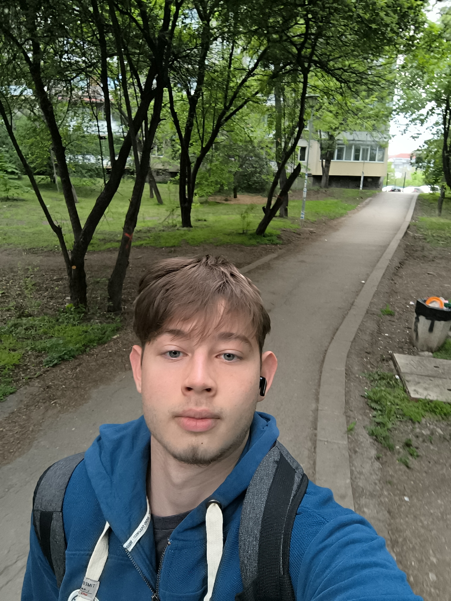 A young man is taking a selfie outdoors. He has short brown hair, blue eyes, and fair skin. He is wearing a blue hooded sweatshirt and a backpack. There is a black wireless earbud in his left ear.
He is standing on a paved pathway in a park-like setting. To his left are several thin trees with green leaves. Behind the trees is a building, possibly a residential apartment complex. The pathway leads uphill and away from the man. To his right, there is a waste bin and some overgrown foliage. The overall impression is that he is walking in a public park or green space in an urban area.
Based on the architecture and vegetation, and given the prompt's mention, it could be located in the city of Sofia, Bulgaria.
