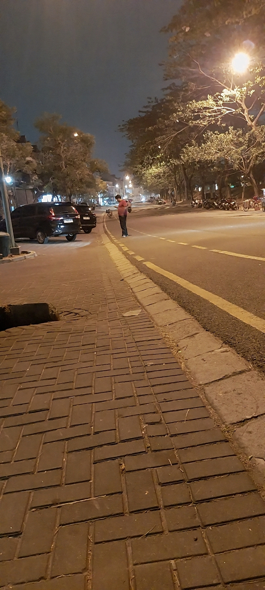 A person in a red shirt and dark pants stands on the side of a road in Kelapa Dua, Indonesia, looking at their phone. The scene is at night, with streetlights illuminating the area. On the left side of the road, there are parked cars, including two black SUVs. The sidewalk is paved with brick-like stones. The road has yellow dividing lines and a concrete curb. Trees line the side of the road, with their branches illuminated by the streetlights. In the background, there are more trees and some buildings. A line of motorcycles is parked on the right side of the road.