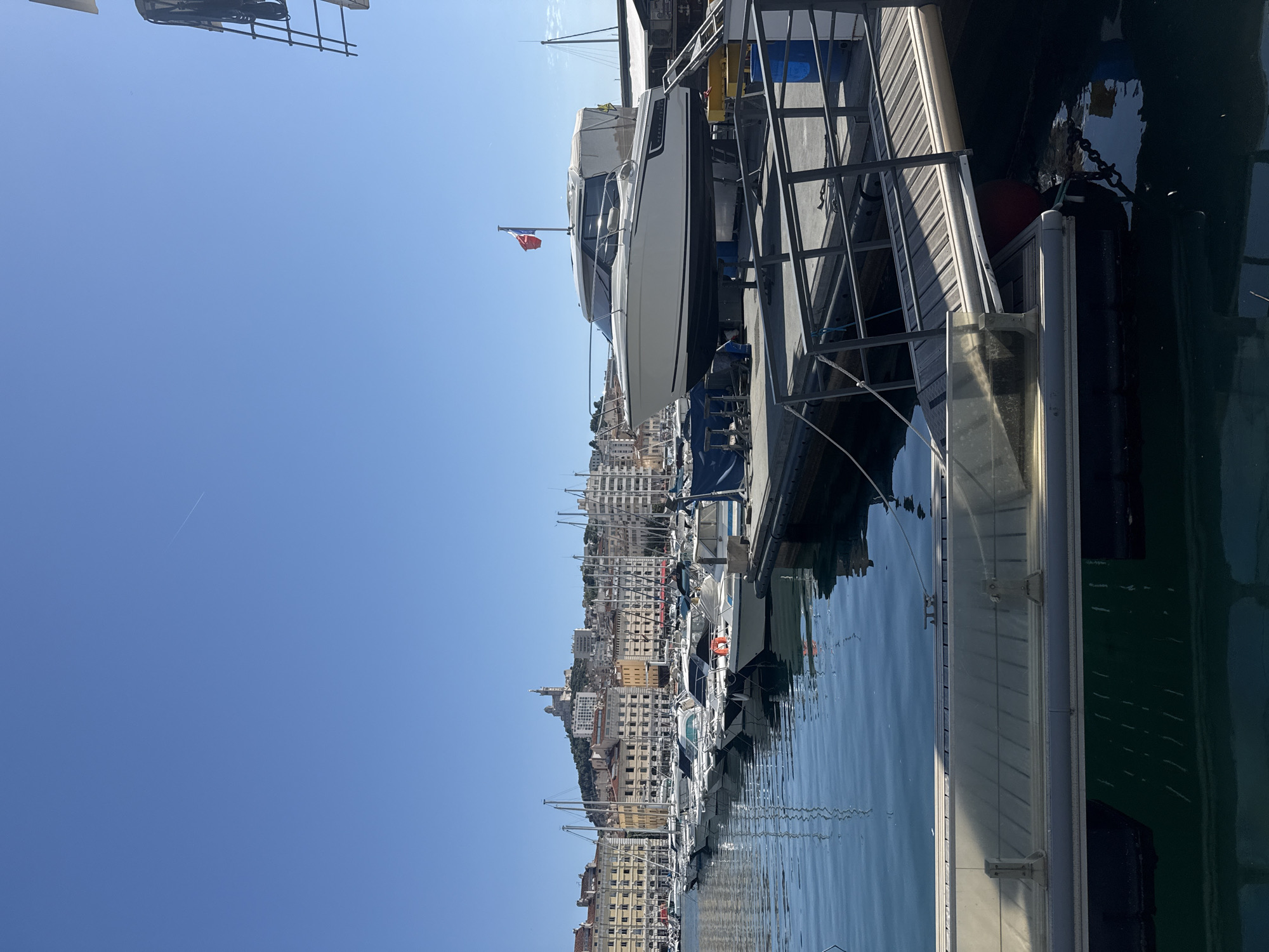 A wide-angle, eye-level shot captures a sunlit marina filled with numerous docked boats and a cityscape in the background under a clear blue sky. The foreground features a section of a large, white boat elevated on a dry dock, with a ramp and metal railings to its left. A French flag waves from the boat's stern. To the left, a line of smaller white boats is moored in the calm, blue water, their reflections shimmering on the surface. Beyond the boats, a dense urban landscape with cream-colored buildings stretches across a hillside, topped by a prominent church or basilica structure. The overall scene is one of a bustling port on a bright, clear day.