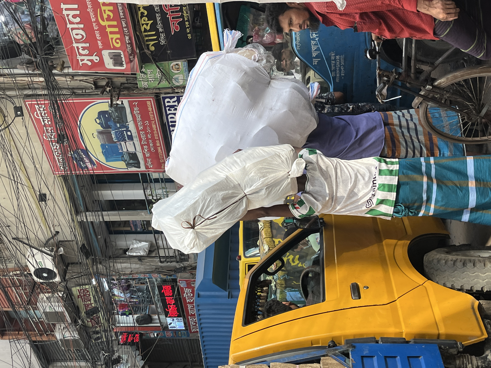 In the bustling city of Dhaka, Bangladesh, a man carries a large, wrapped white package on his shoulders, likely headed for a yellow truck. The man wears a white t-shirt with green accents, a blue and white striped lungi (traditional garment), and seems to be engaged in manual labor. The truck, with a blue flatbed, is situated amidst a congested street filled with overhead electrical wires and colorful, densely packed signage. The signs in Bengali advertise various businesses, including a luggage shop and a bag wholesaler. A man in a red shirt can be seen nearby, possibly involved in the same activity or transportation. The scene showcases the typical, chaotic, and industrious environment often found in Dhaka.