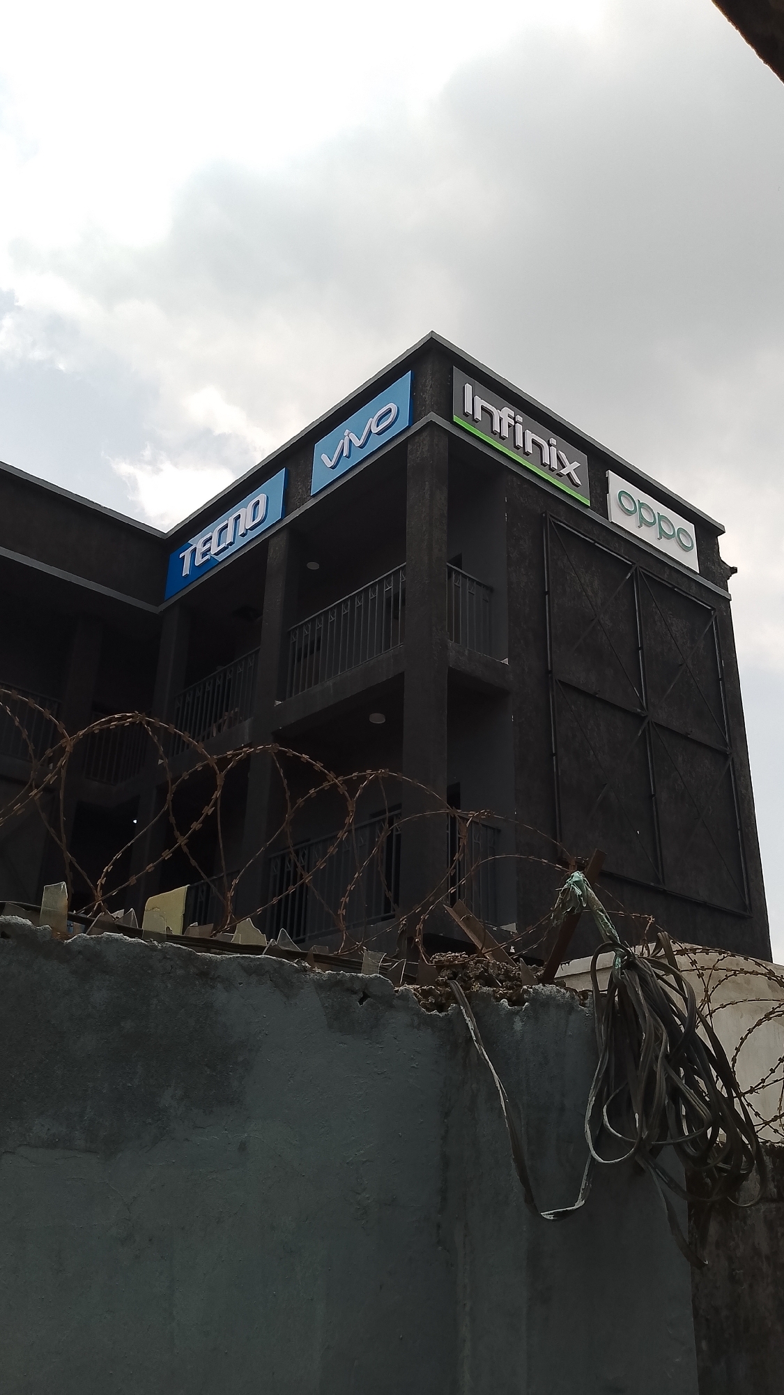 A two-story dark-colored building stands behind a weathered, grey concrete wall topped with barbed wire, a stark contrast to the cloudy sky above. The building's facade is marked with the logos of several mobile phone brands: TECNO, VIVO, Infinix, and OPPO. The structure features iron bars acting as balconies. An assortment of cables hangs tangled near the barbed wire, adding to the unkempt appearance of the perimeter. The location is likely Effurun, Nigeria, based on the presence of these commonly advertised mobile phone brands and the architectural style of the building. No people are visible in the scene.