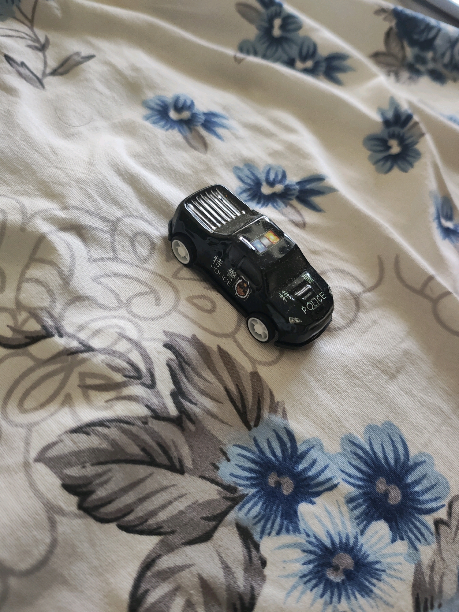 A close-up, overhead shot captures a black toy police car resting on a white fabric with a floral pattern. The fabric features blue and gray flowers with dark green leaves. The police car is positioned diagonally, with its front facing the bottom right of the frame. It has white wheels and displays the word "POLICE" on its side, along with some Chinese characters and a police emblem. The roof of the car has a ribbed detail and what appears to be flashing lights in blue and orange. The scene is well-lit, with soft shadows indicating an indoor setting. There are no people or animals visible in the image. The overall impression is that of a child's toy on a bed or sofa. The image does not contain any information or visual cues to suggest its location within the city of Jaipur Municipal Corporation, India.