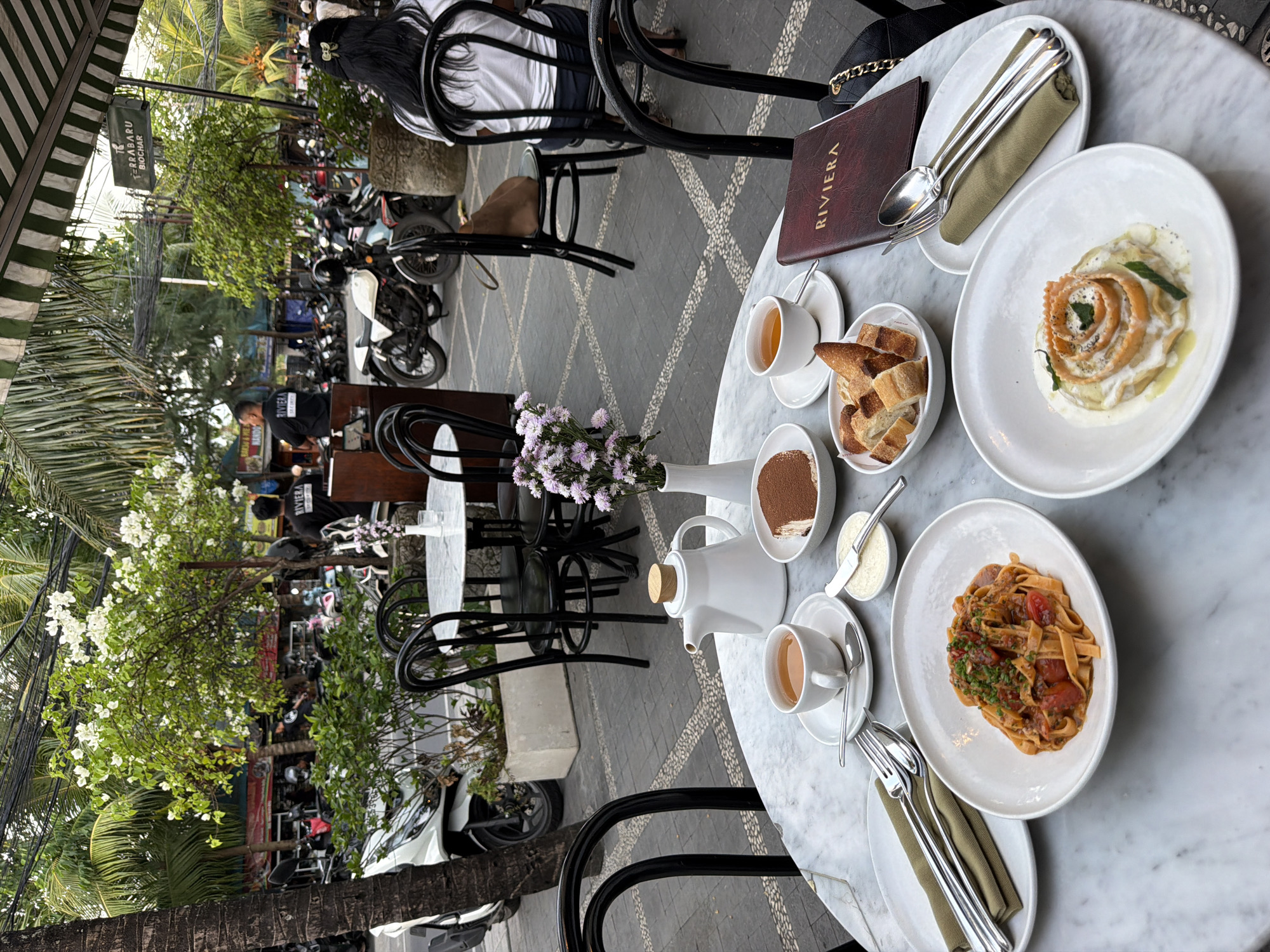 A wide shot captures an outdoor dining scene at the Riviera restaurant in North Kuta, Indonesia. The image is dominated by a marble-topped table laden with food and drinks, set against a backdrop of a bustling street. The table features two plates of pasta, a bowl of bread, a slice of tiramisu, a cup of tea, and a teapot. The restaurant's menu, a dark red booklet, rests near a set of cutlery. Black metal chairs surround the table, with one occupied by a woman with long dark hair wearing a white shirt.
In the background, motorcycles are parked along the street, and several people are visible, including two men wearing black t-shirts with "RIVIERA" printed on them. Lush green trees and white flowering plants adorn the scene, suggesting a tropical ambiance. A striped awning of green and white hangs overhead, providing shade. The overall impression is of a vibrant, lively dining experience in a tropical urban setting.