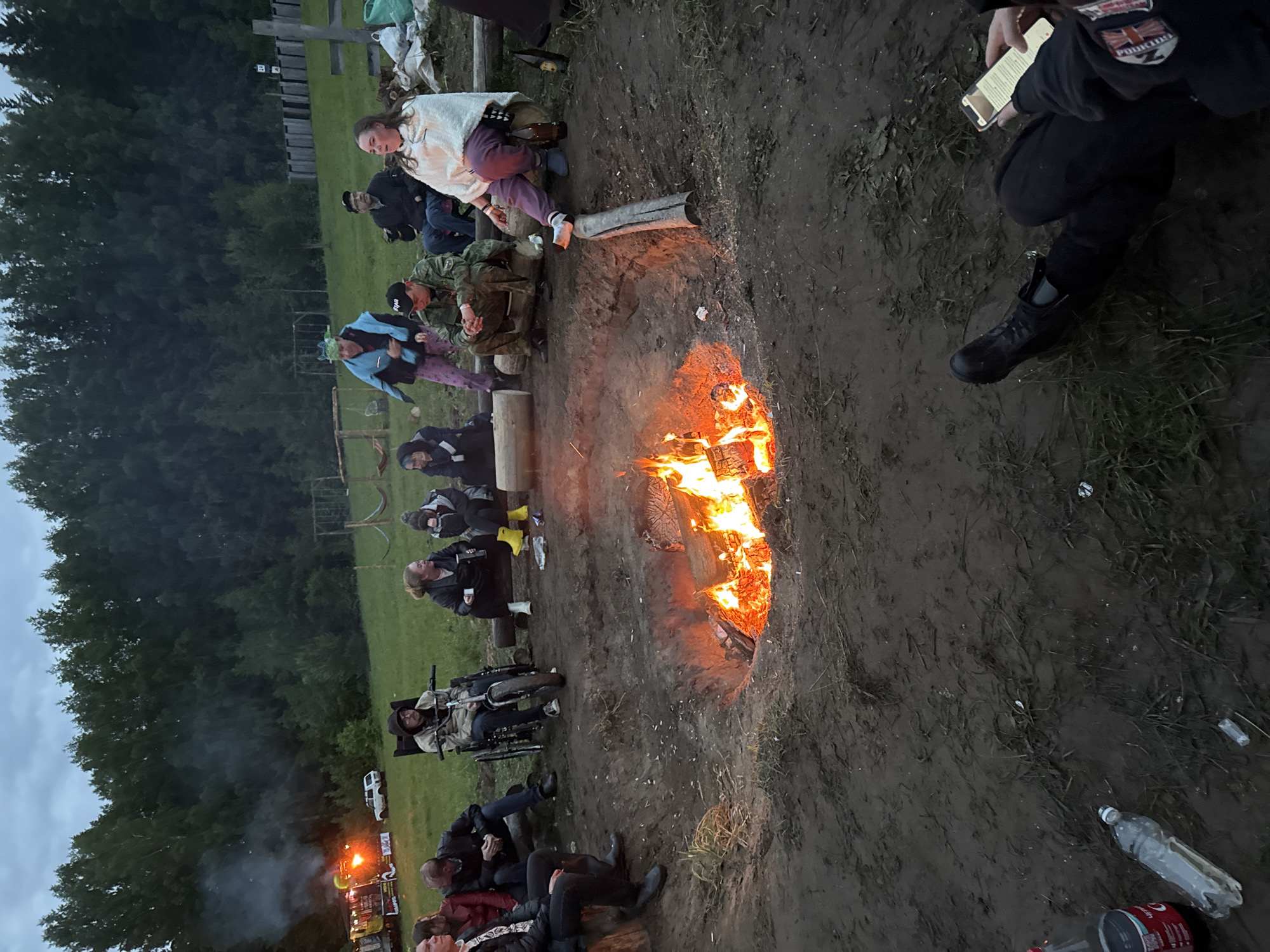 The scene depicts an outdoor gathering around a large bonfire pit in a muddy field located in Большая Кивара, Russia, during dusk or early night. Approximately fourteen individuals are visible, primarily seated on logs arranged around the central fire, which contains burning wood and emits significant light and some smoke. One person is seated in a specialized wheelchair with a large front wheel, positioned on the left side of the fire.
The attendees are dressed in various casual and outdoor attire, including jackets, hoodies, and one person wearing military-style camouflage. Several individuals are holding drinks, including a visible Coca-Cola bottle, a clear plastic water bottle in the foreground, and mugs held by others. An empty beer bottle rests on the log next to a woman wrapped in a white shawl, who is pointing towards the fire. In the immediate foreground, a person's legs and right arm are visible, holding a smartphone with the screen illuminated.
The environment is characterized by damp, muddy ground with sparse grass. In the background to the left, a large, illuminated vehicle, possibly a truck or recreational vehicle, with visible smoke, is parked behind a smaller white car. To the right background, a dense forest lines the field, with a wooden fence, a signpost, and rustic wooden playground structures visible. The sky is overcast, contributing to the muted natural light.
