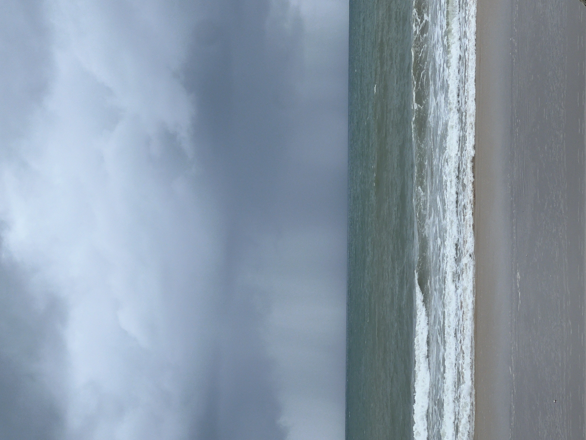 The image depicts a beach scene under an overcast sky. The left portion of the frame is filled with grey, cloudy sky, while the right portion shows the ocean meeting the shore. Waves are visible crashing onto a light-colored sandy beach. No people are visible in the image. The location is Unknown, Unknown.