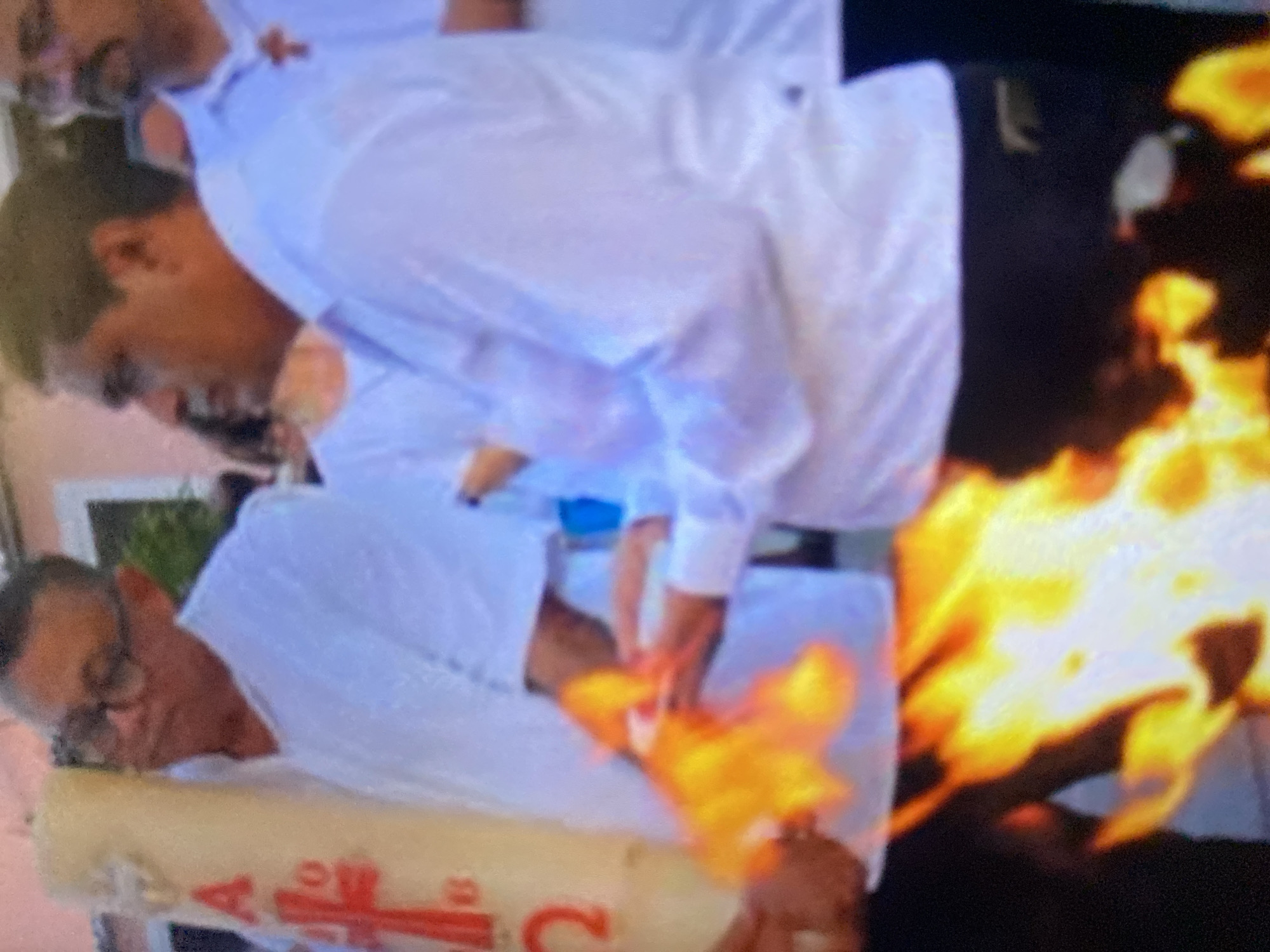 The image depicts two men dressed in white shirts and dark pants, appearing to be clergymen or religious figures, standing next to a large candle. The candle is decorated with a red cross and lettering, and the top of it is actively burning, with a visible flame emanating upwards. The location is Tinaquillo, Venezuela.