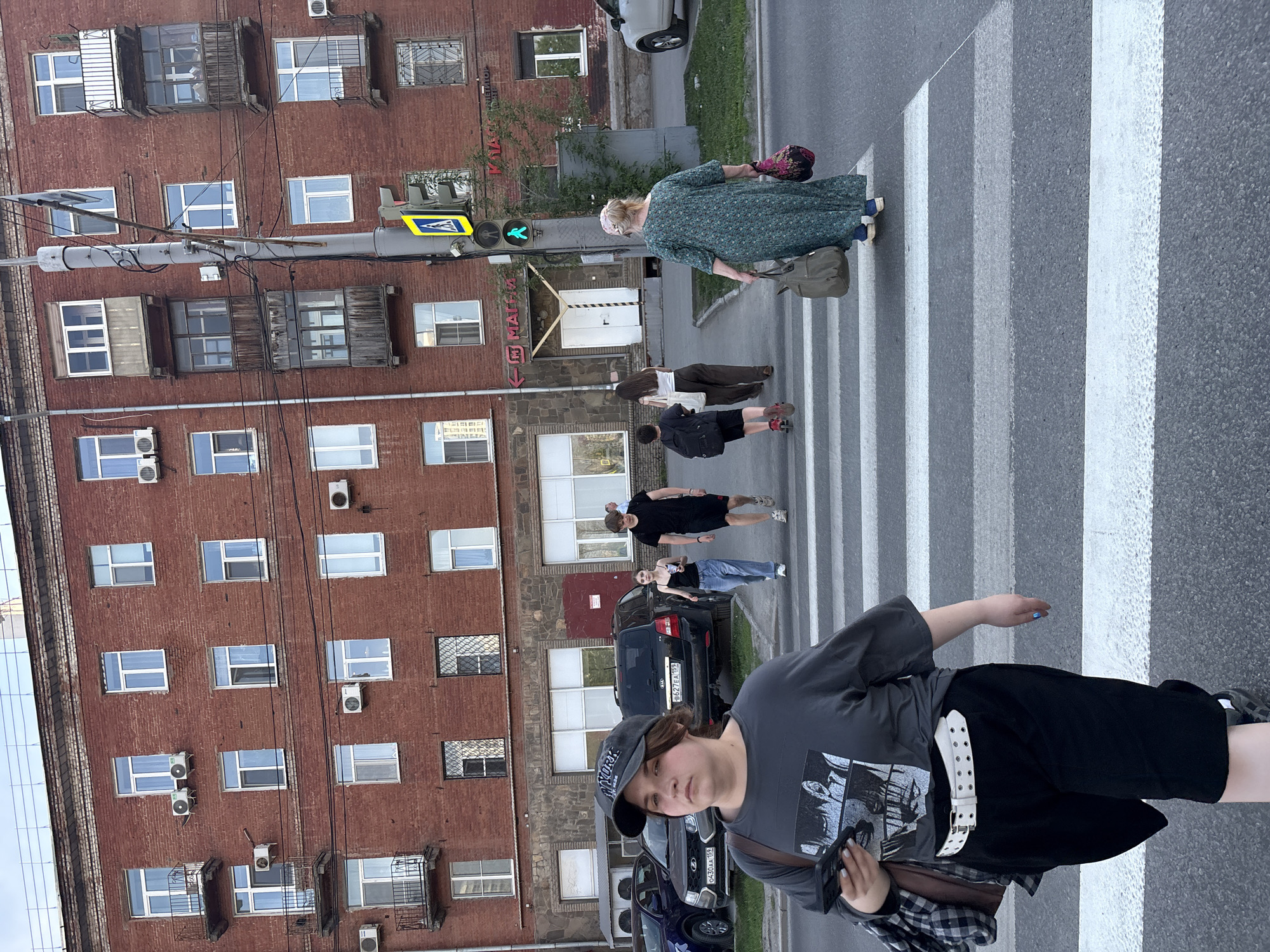 The image depicts a street scene in Perm, Russia, with pedestrians crossing a zebra-striped crosswalk in front of a red brick apartment building. The building has multiple rows of windows, some with small air conditioning units. Overhead electrical cables are visible. A green traffic light for pedestrians is illuminated. People are crossing the street, including an older woman in a green dress with a bag and hat, as well as several younger individuals. Cars are parked along the side of the street. A person in the foreground is walking away from the camera, wearing a t-shirt, shorts, and a baseball cap, with a plaid shirt tied around their waist.