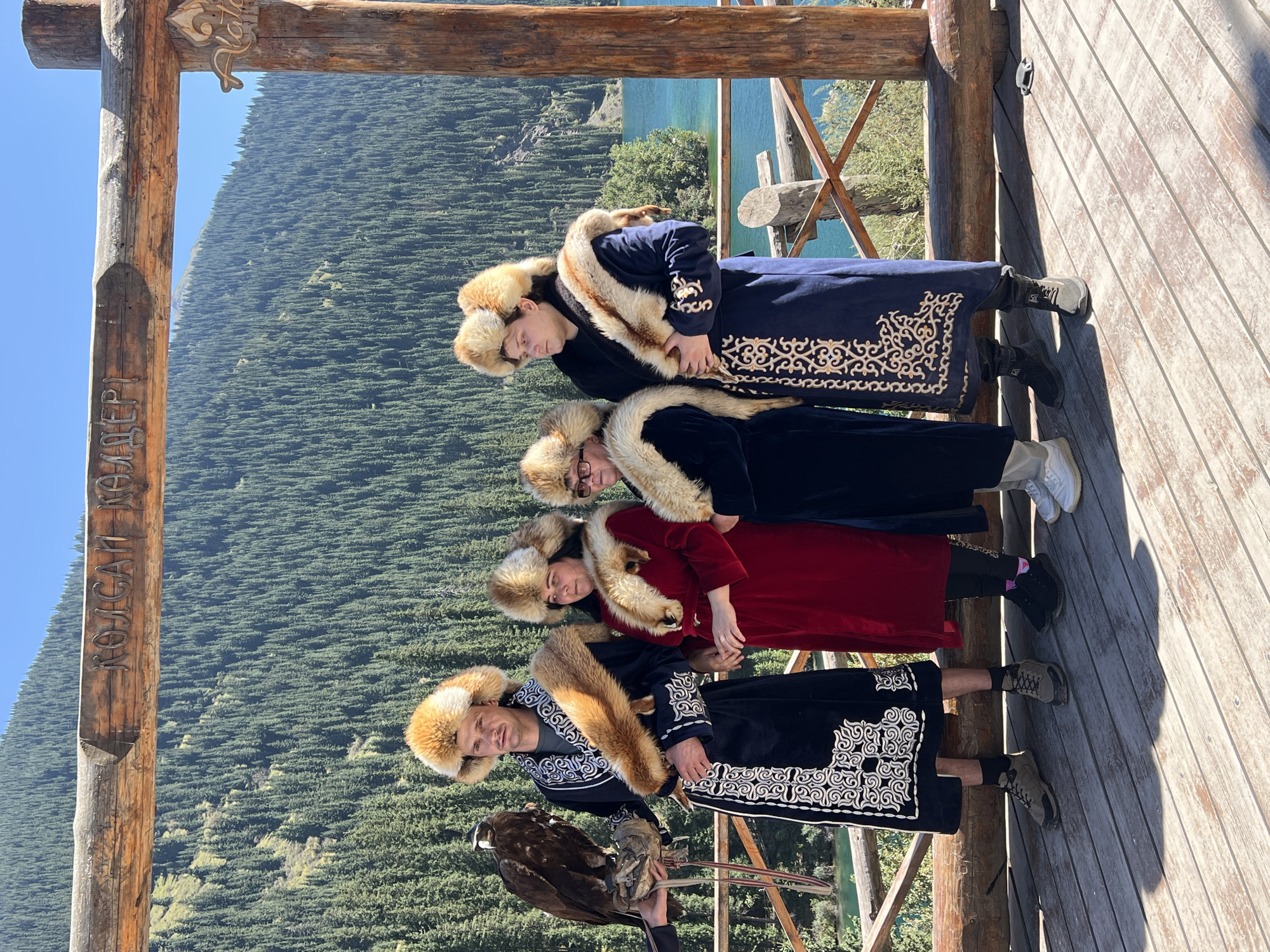 In Kurmeti, Kazakhstan, four individuals are posing for a photograph on a wooden platform, with a scenic backdrop of a mountain range covered in dense evergreen trees and a clear blue lake. The individuals are dressed in traditional attire, featuring elaborate fur-lined hats and cloaks.
From left to right:
The first individual, a male, is holding a golden eagle on his gloved left hand. He is wearing a black, long-sleeved cloak adorned with white, intricate embroidery on the hem and sleeves. His hat is a large, fluffy, orange-brown fur hat. He is also wearing dark pants and light brown hiking boots.
The second individual, a female, is wearing a vibrant red, long-sleeved dress and a matching orange-brown fur hat. She has a fur stole around her neck. She is wearing black leggings and black athletic shoes.
The third individual, also appearing to be female, is wearing a dark blue, long-sleeved cloak with fur trim around the collar and a fur hat. She has glasses and appears to be wearing dark pants and white athletic shoes.
The fourth individual, a male, is wearing a dark blue, long-sleeved cloak with gold embroidery on the hem and sleeves. His hat is a large, fluffy, orange-brown fur hat, similar to the first individual's. He is wearing dark pants and dark hiking boots.
A wooden archway, with carved lettering in Cyrillic script, spans above the individuals. The environment suggests a tourist or cultural attraction, likely in a natural, mountainous area. The lighting indicates a sunny day.