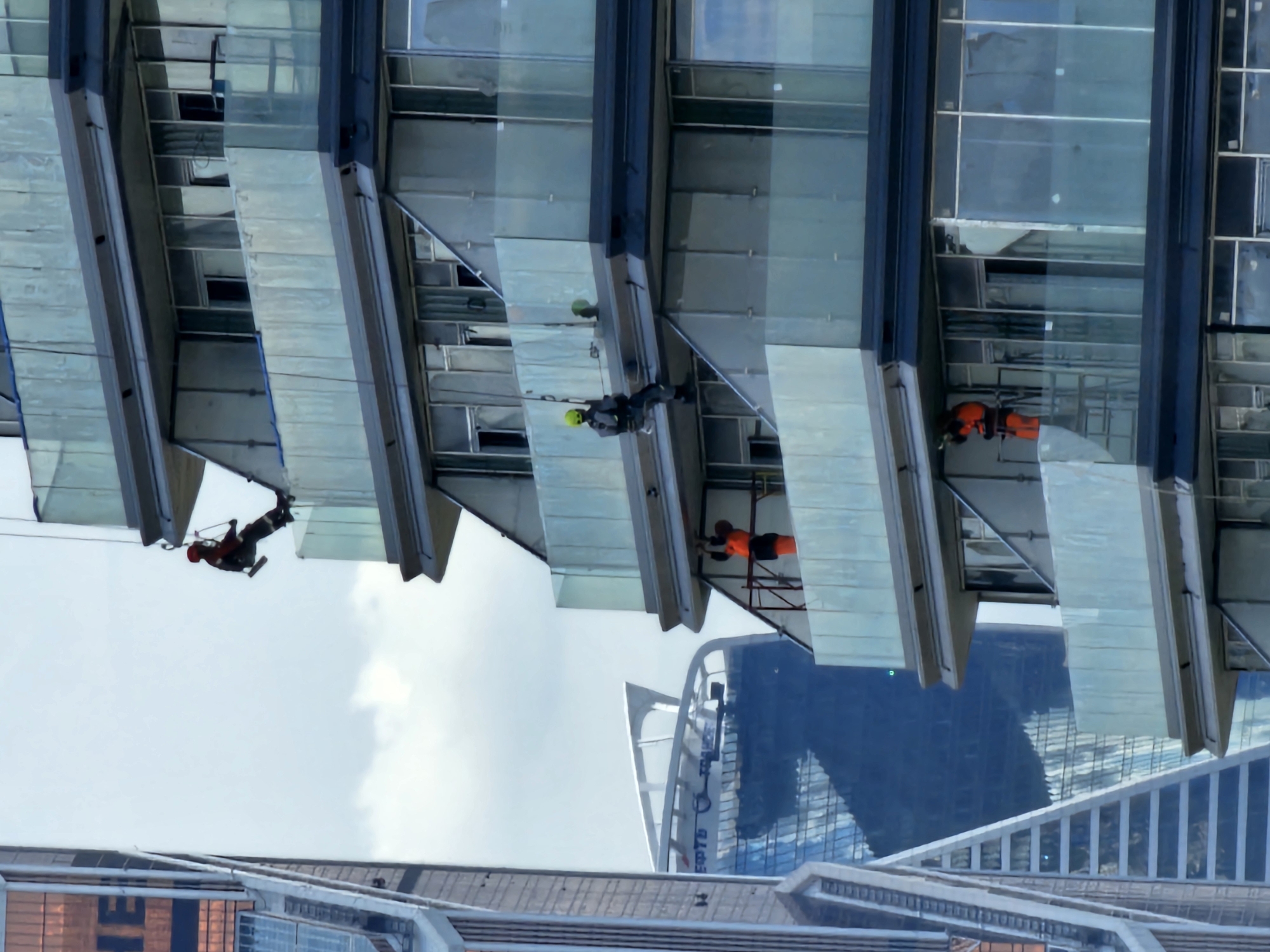 In Moscow, Russia, the image captures construction workers engaged in work on a modern building with a facade of glass and metal. The building features multiple levels with balconies, each enclosed by glass railings. In the foreground, three construction workers are visible. The uppermost worker, dressed in a red helmet and jacket, is suspended from above, appearing to be rappelling down the side of the building. Below this worker, another individual in a green helmet and dark attire is positioned on a balcony, also seemingly engaged in work. Further down, a third worker, wearing an orange vest and trousers, is standing on a balcony, possibly preparing to ascend or descend. In the background, other skyscrapers contribute to the urban landscape, with one prominent building exhibiting a mirrored facade reflecting the sky and surrounding structures. The sky is overcast, suggesting a cloudy day.