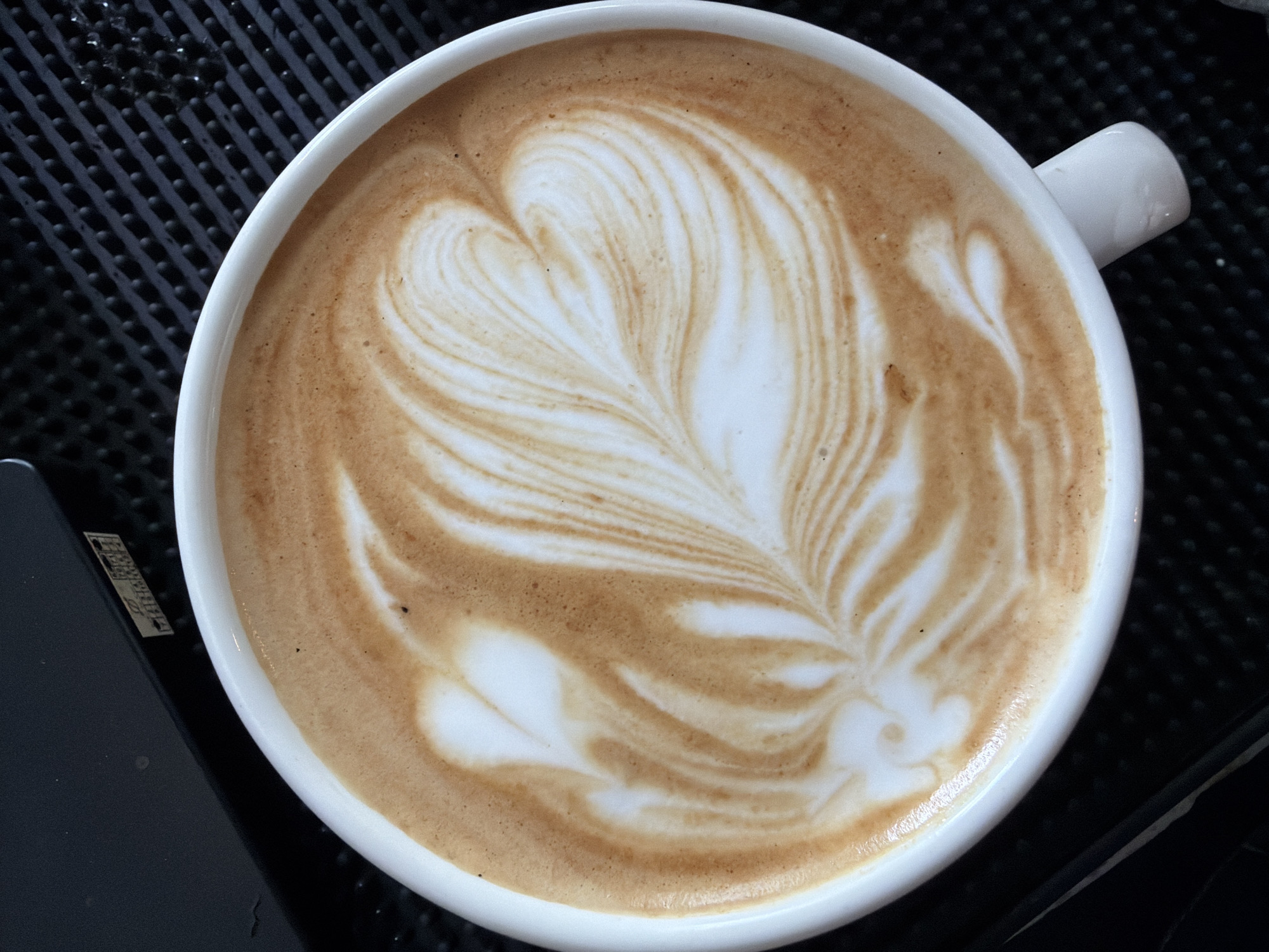 A close-up, overhead shot captures a white ceramic mug of latte art, its foam meticulously patterned with a swirling leaf design in shades of cream and light brown. The mug rests on a black textured surface, possibly a bar counter with a grid pattern. In the upper left corner, a sliver of a dark, flat object, potentially a machine or counter edge, is visible. A small sticker with a series of numbers and symbols is affixed to this dark object. The overall scene suggests a coffee shop environment, possibly in Karon, Thailand, where such artistic beverage presentations are common.