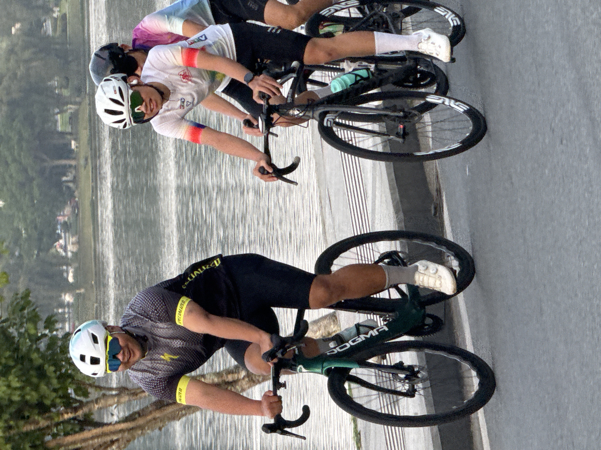 Three cyclists, two men and one person whose gender is not discernible, are riding their bicycles on a paved path next to Xuân Hương Lake in Đà Lạt, Vietnam. The cyclist in the foreground on the left is a man wearing a white and blue helmet, yellow and black cycling jersey, and black cycling shorts. He is on a dark green bicycle. To his right, another cyclist, a man, is wearing a white helmet, white cycling jersey with colorful accents on the sleeves, and black cycling shorts. He is on a black bicycle. Behind him, another cyclist, whose gender is not discernible, is wearing a black helmet and a tie-dye colored cycling jersey. The cyclists are all wearing cycling shoes and sunglasses. The lake is visible in the background, with trees and a bridge also present. The sky appears overcast. The image is taken from a slightly low angle, focusing on the cyclists and their bicycles.