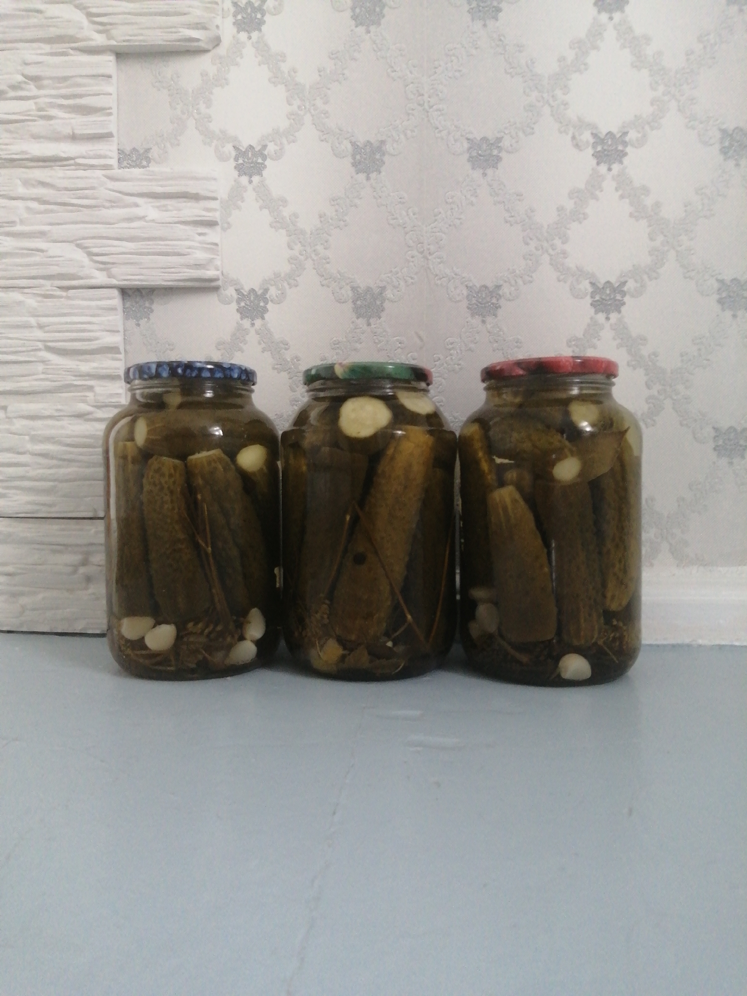 Three glass jars filled with pickled cucumbers and garlic are arranged in a row on a light blue surface. The jars are partially obscured by the jars in front of them. Behind the jars is a wall with a textured white stone-like pattern on the left and a white patterned wallpaper on the right. The scene appears to be indoors, likely in a residential setting in Slonim, Belarus. No people are visible in the image, and no specific actions or interactions are depicted.